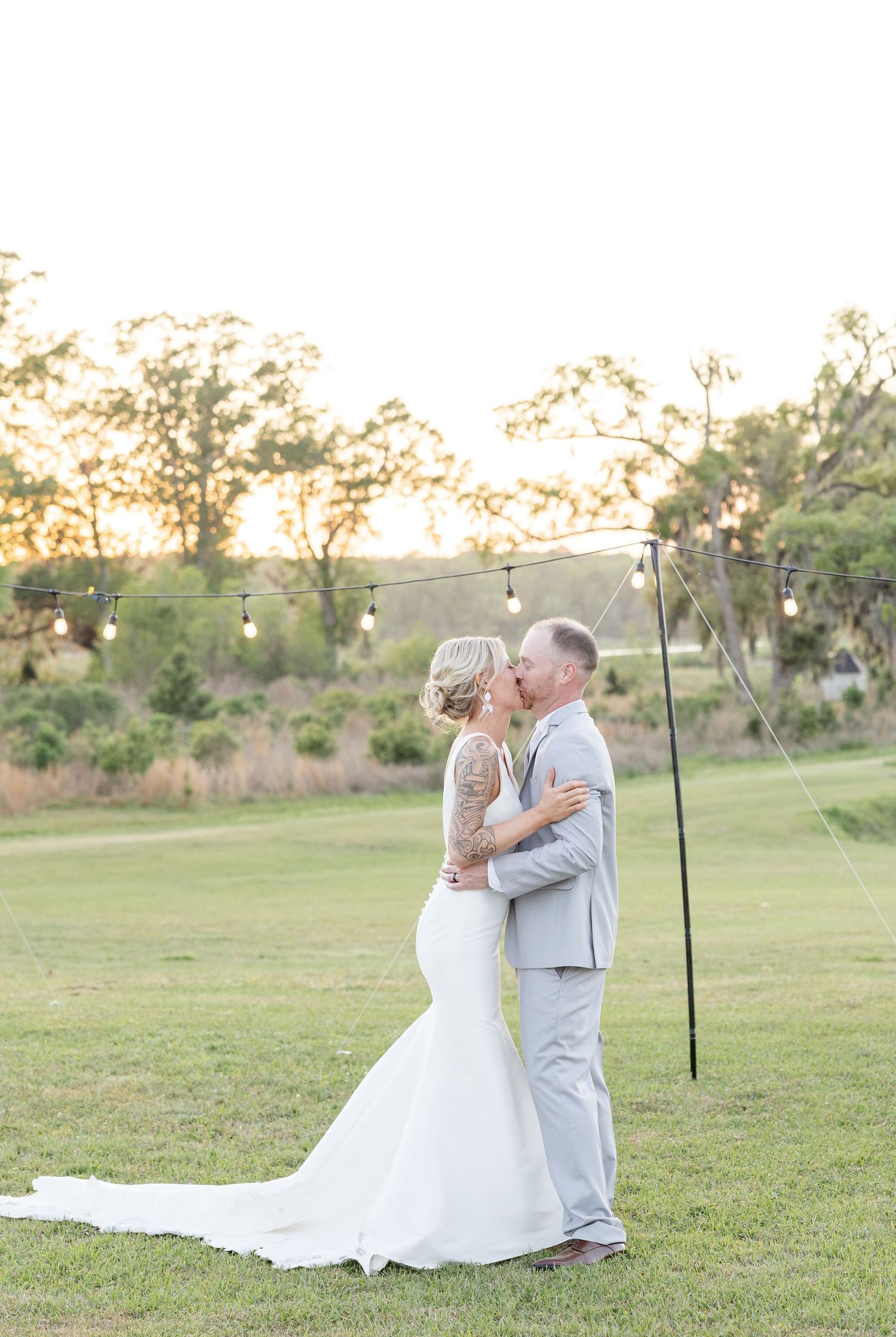 Bride and groom share a kiss at sunset under string lights at an outdoor wedding ceremony.