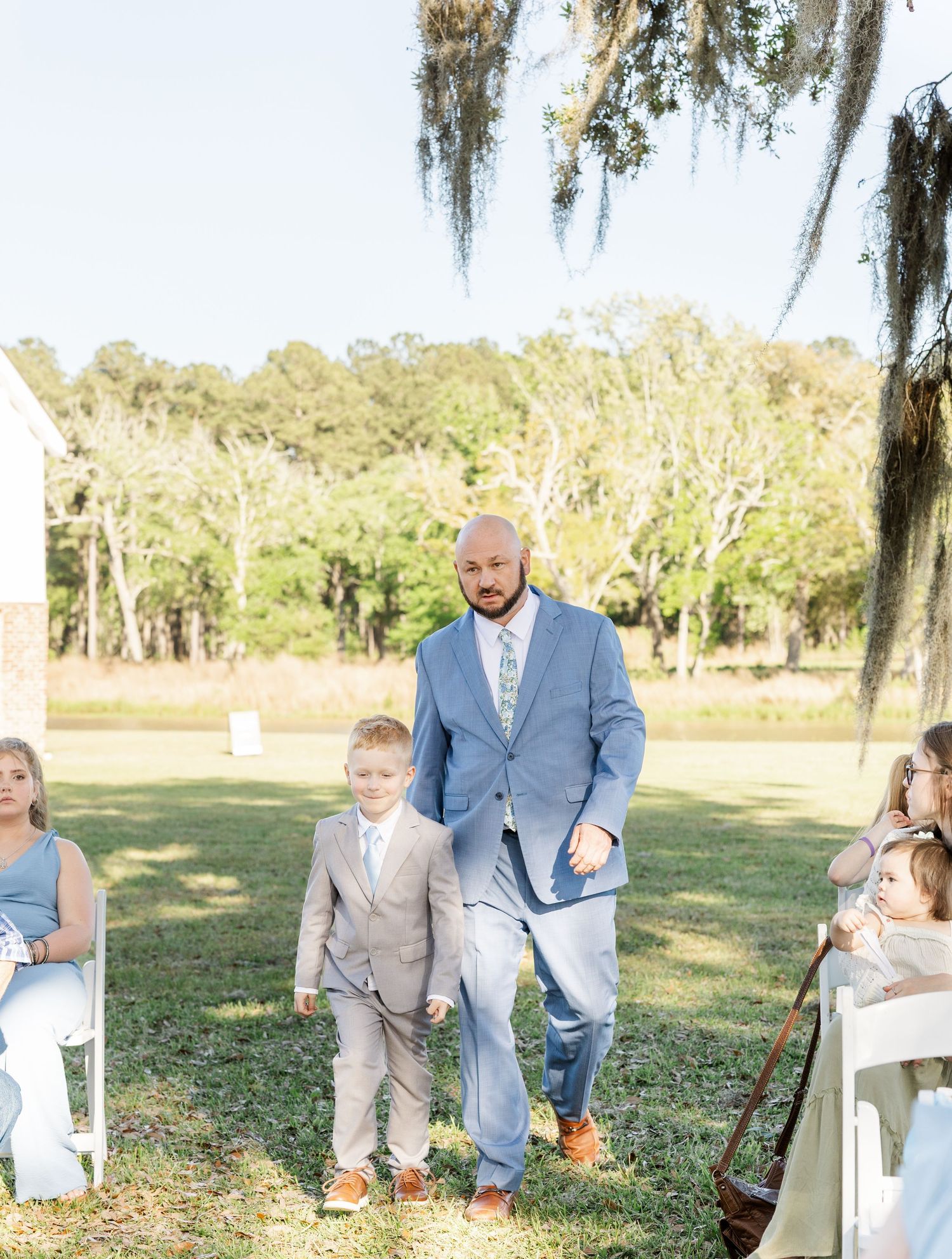 Groom in blue suit walking young ring bearer down the aisle at outdoor wedding ceremony.