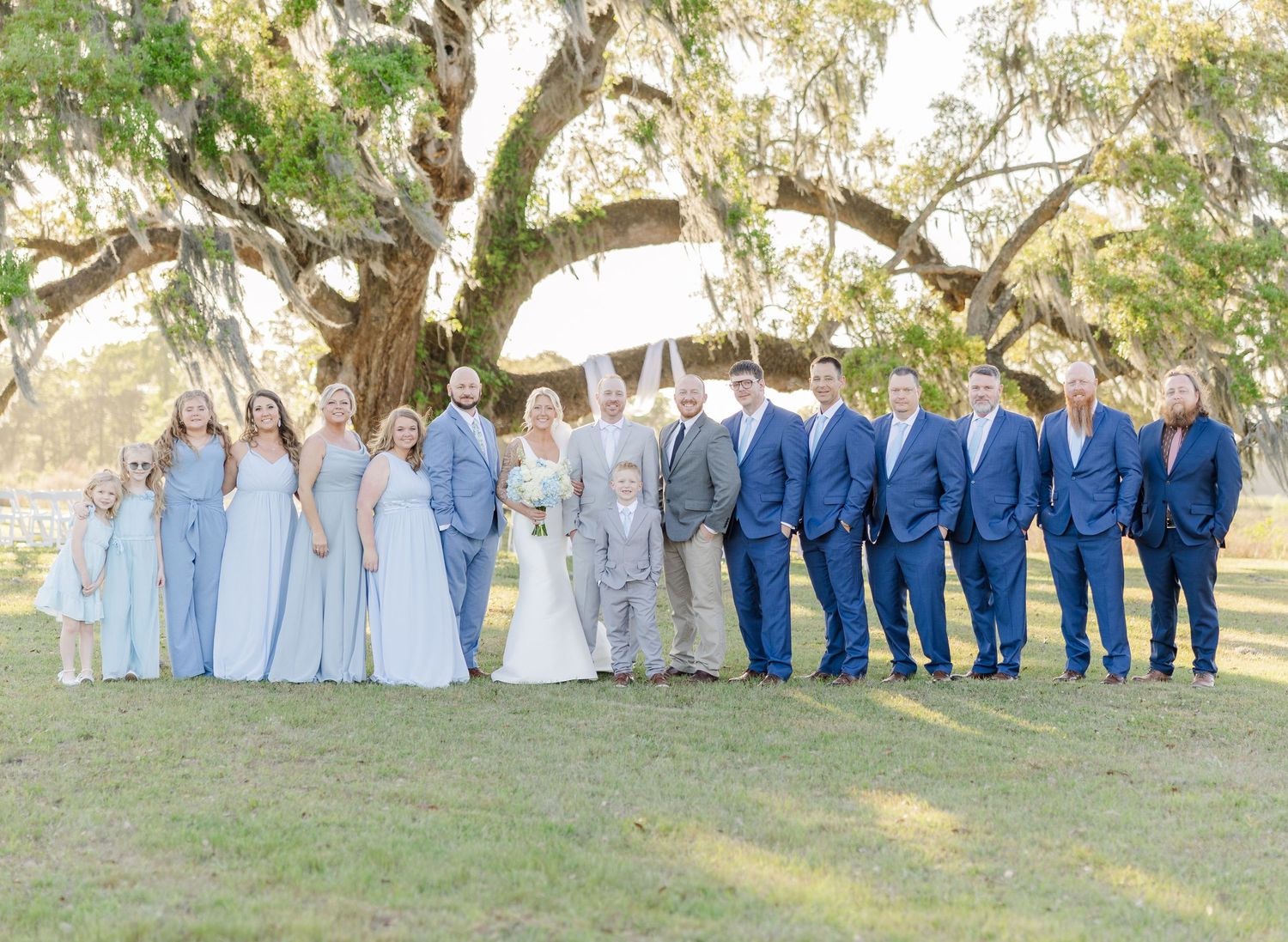 Large wedding party posing under a sprawling oak tree, bridesmaids in blue gowns, groomsmen in navy.
