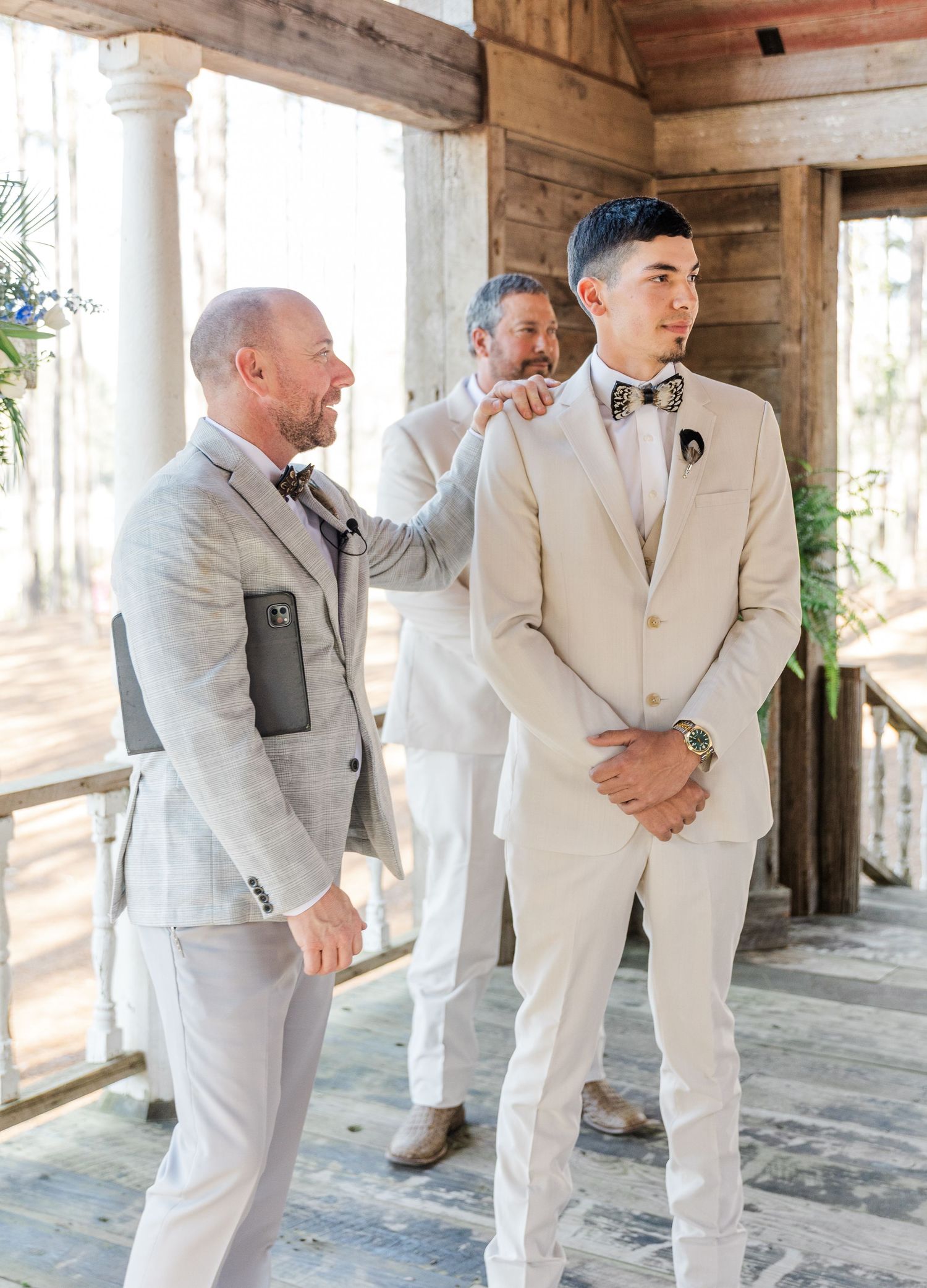 Groomsmen helping groom adjust his outfit inside a rustic wooden wedding venue pavilion.