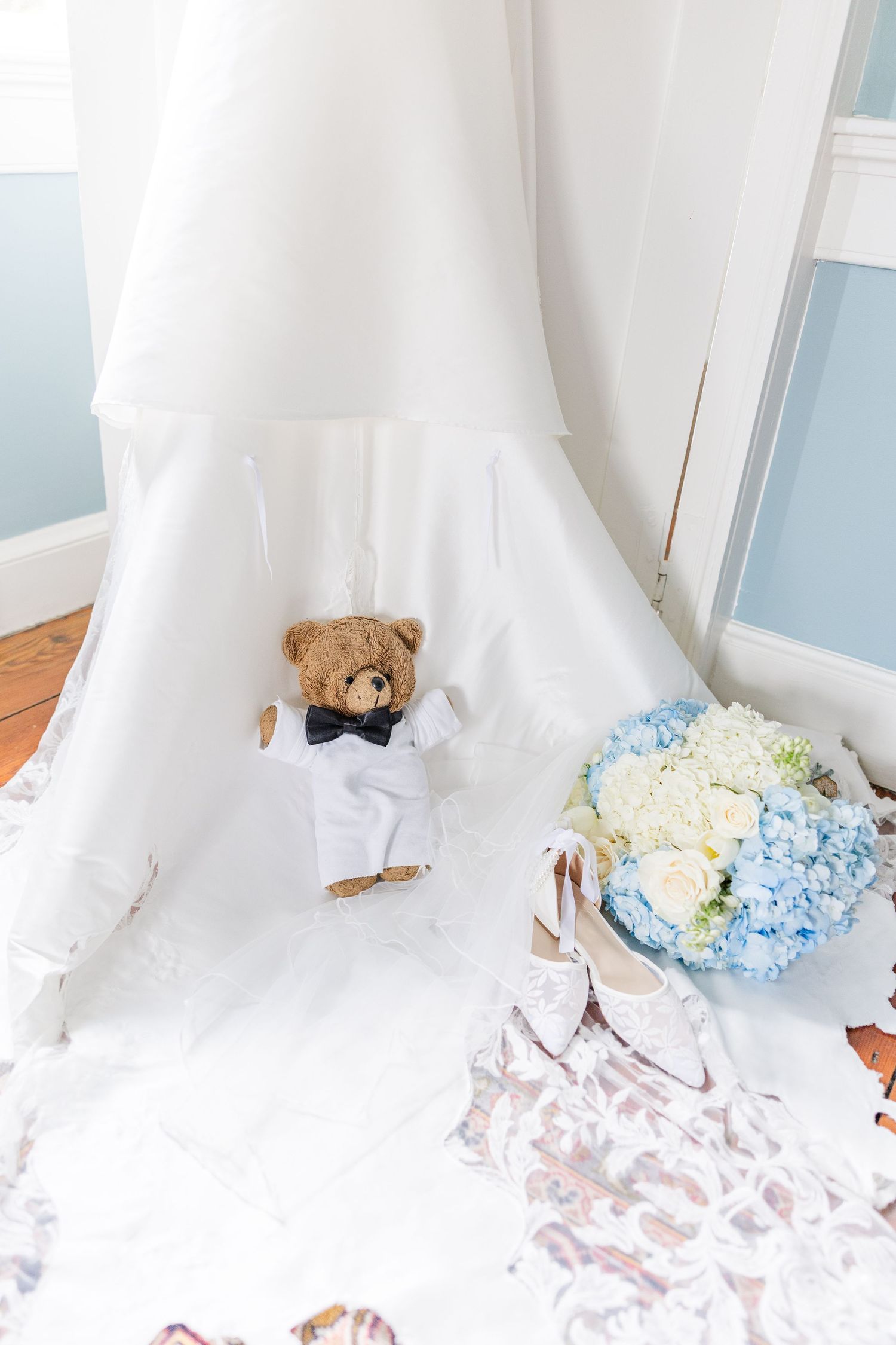 Teddy bear in tuxedo sits beside bridal bouquet of blue hydrangeas and wedding shoes under gown.