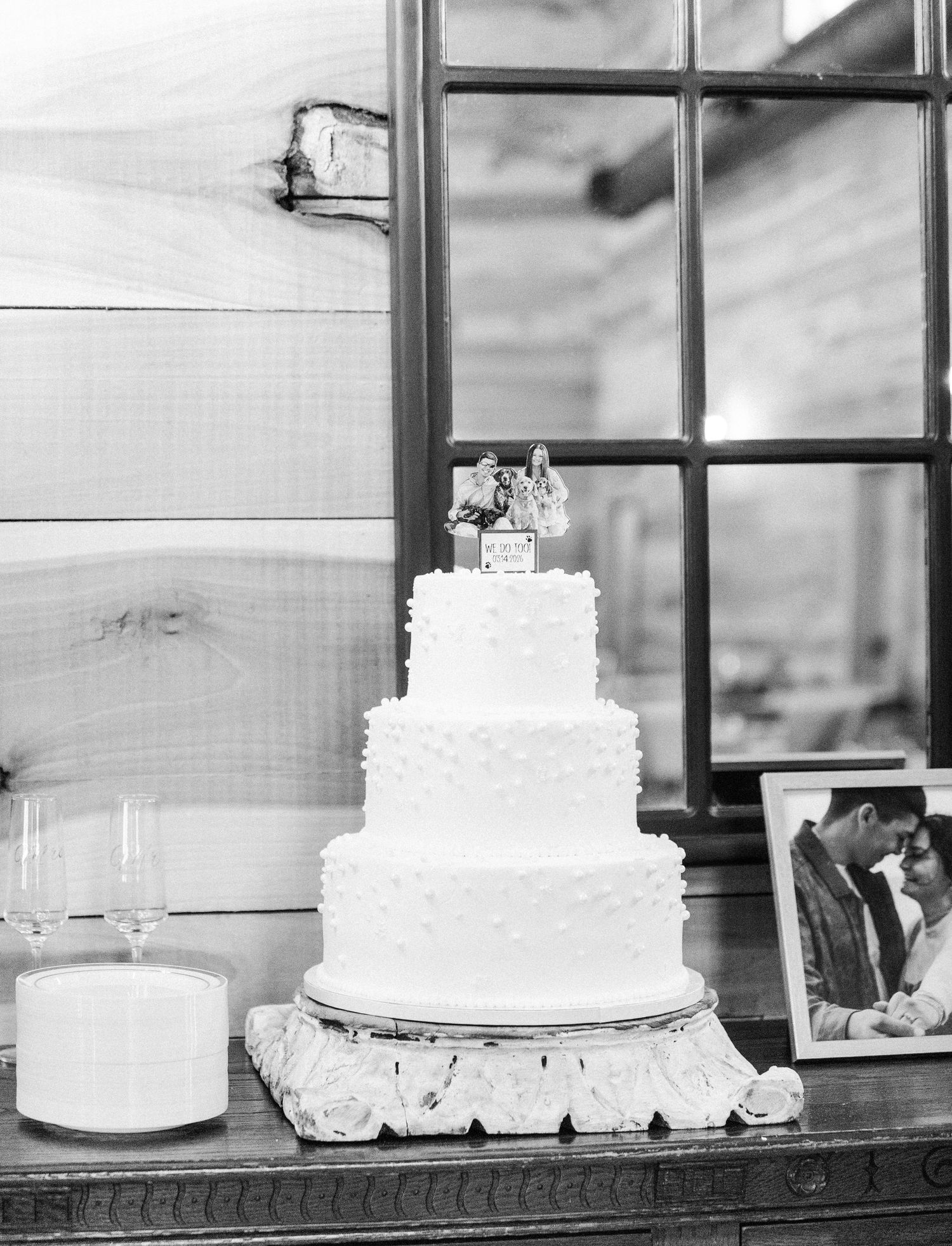 Classic three-tier white rosette wedding cake with custom topper displayed on ornate stand in black and white photo.