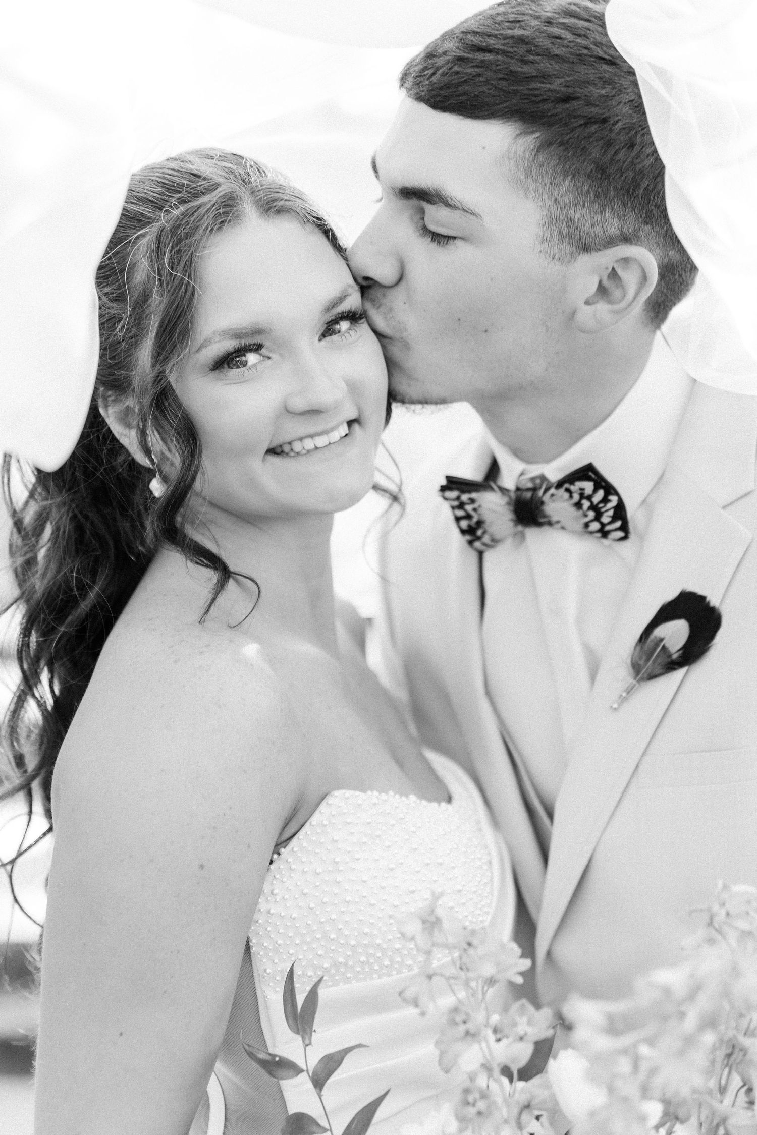 Black and white photo of groom kissing bride on cheek as she smiles holding a bouquet.