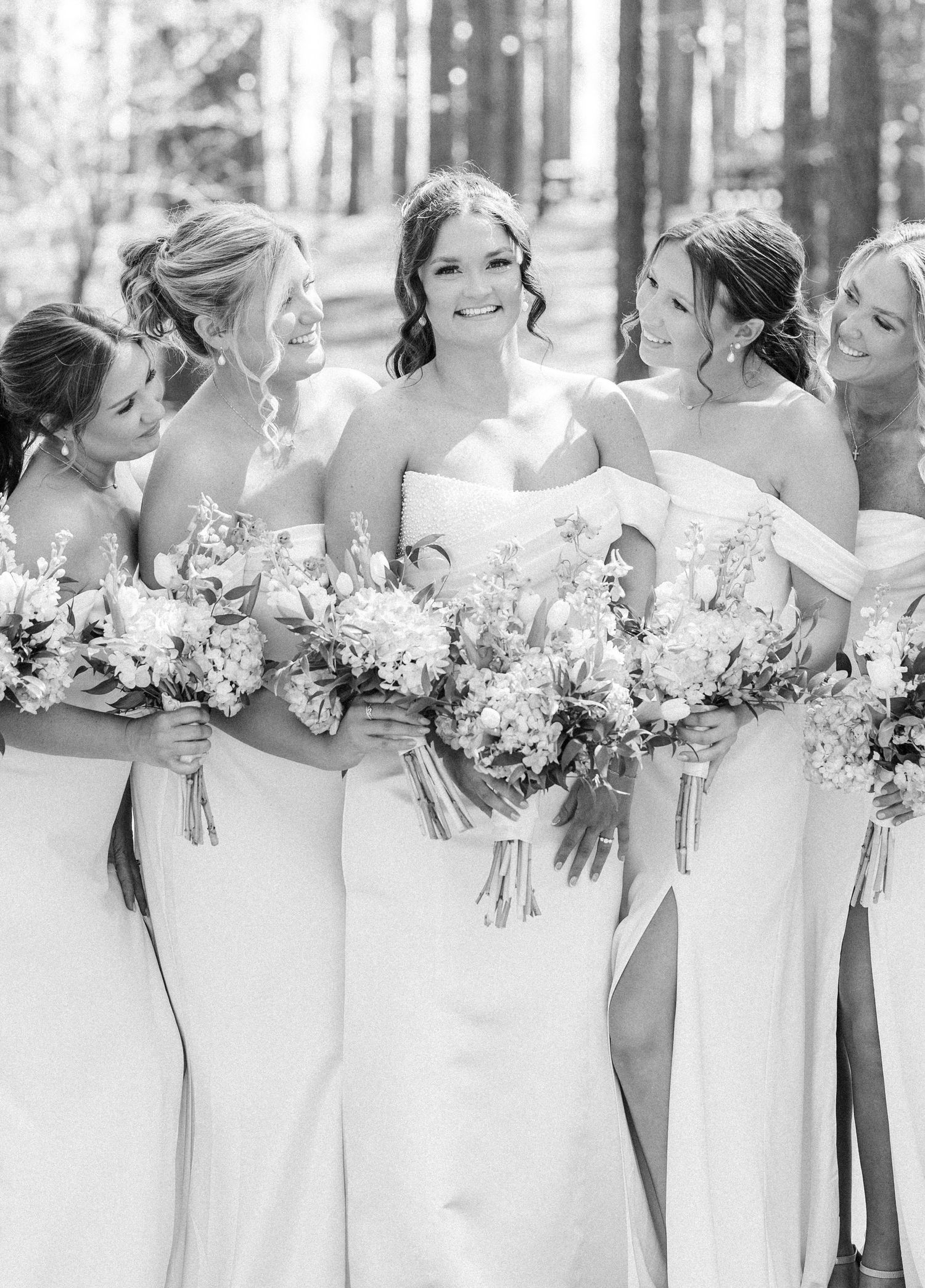 Black and white photo of bride surrounded by smiling bridesmaids holding floral bouquets in pine forest.