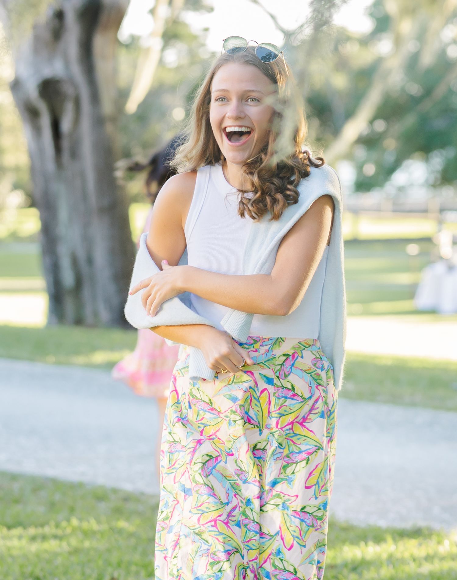 A joyful figure in a white sleeveless top and colorful patterned pants stands outdoors on a sunny day.