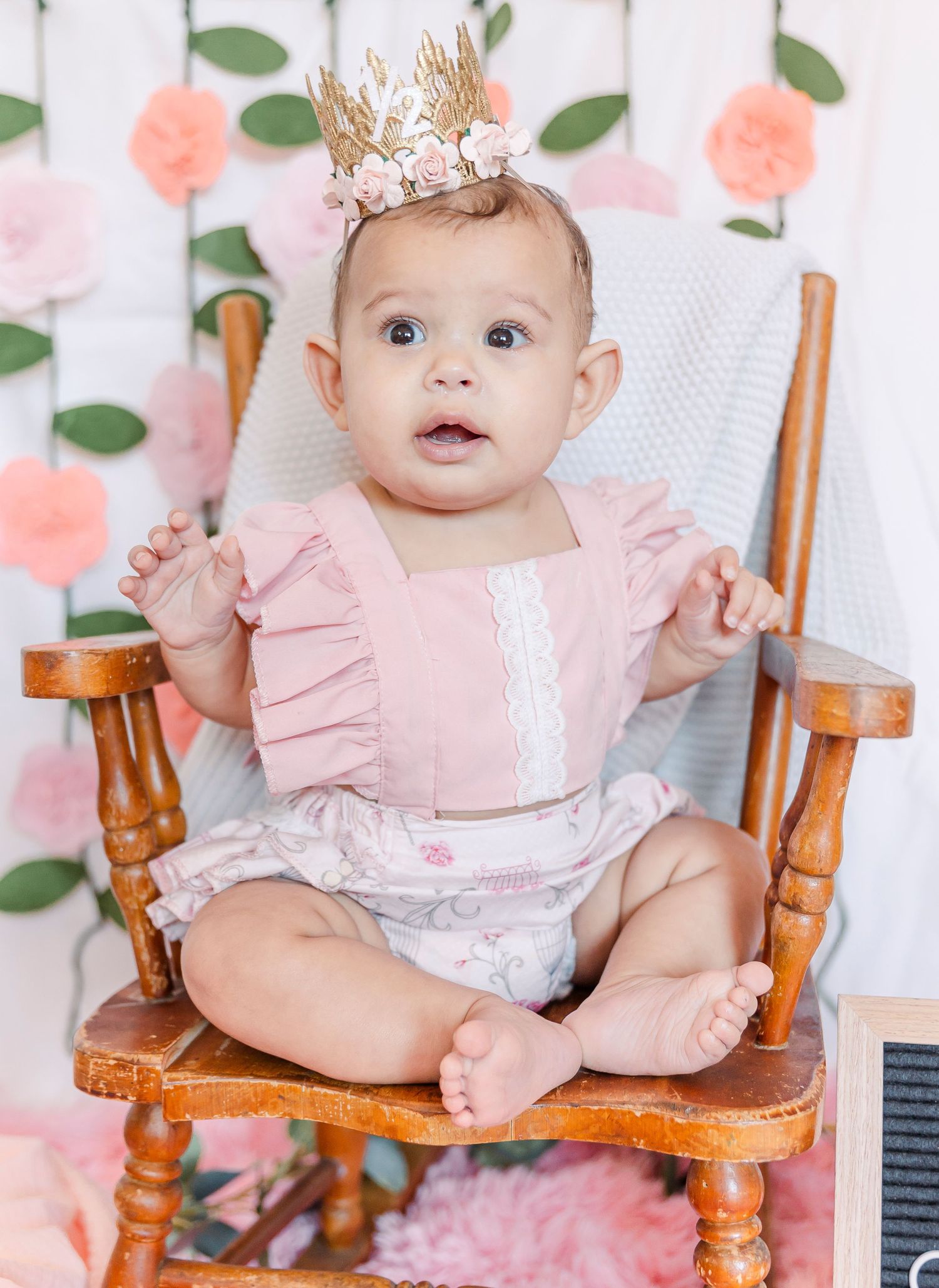 Milestone photo featuring ruffled pink dress and gold crown on wooden rocking chair.