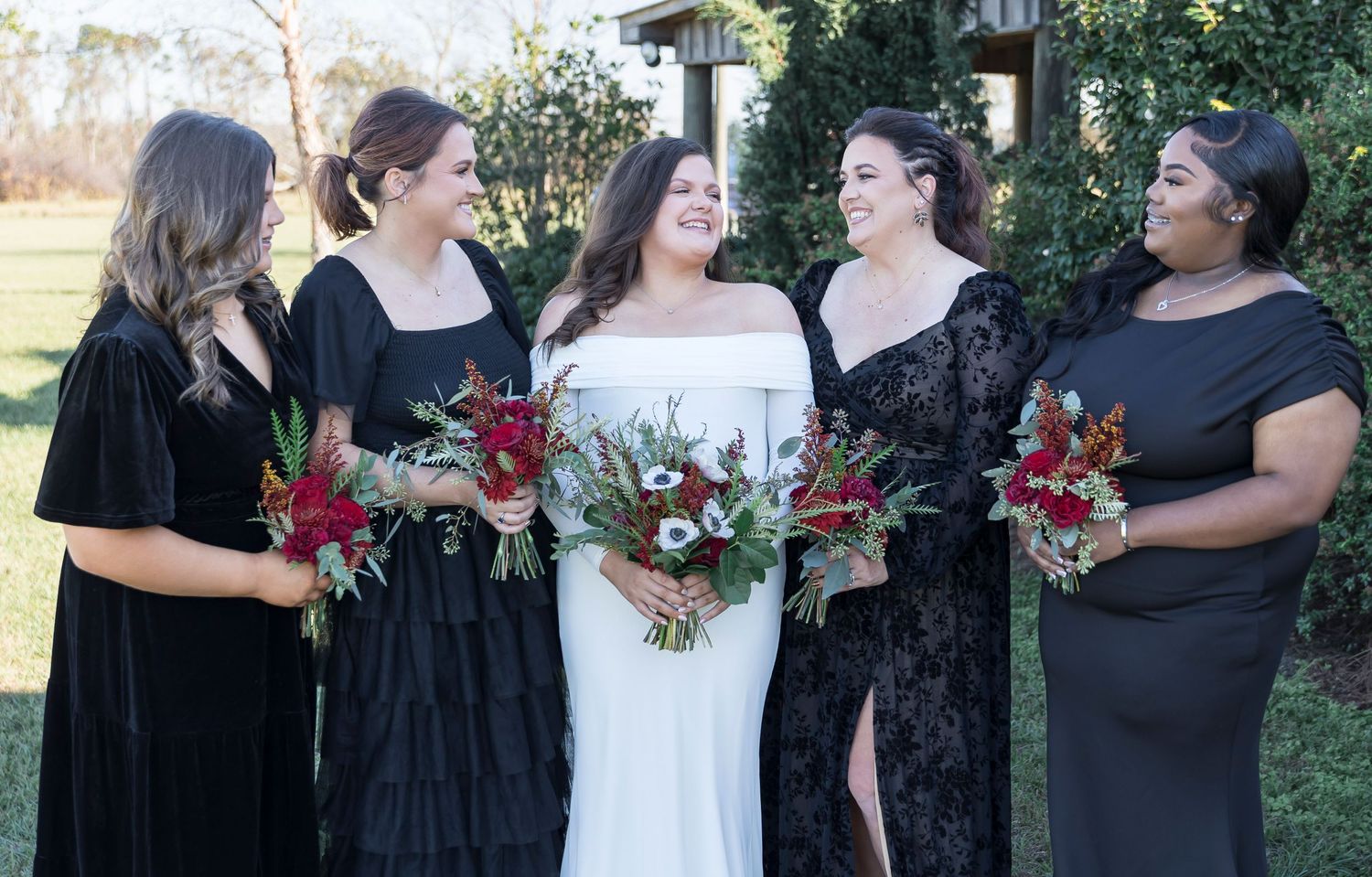 Bridal party in elegant black dresses holds vibrant red and white bouquets against outdoor garden setting.