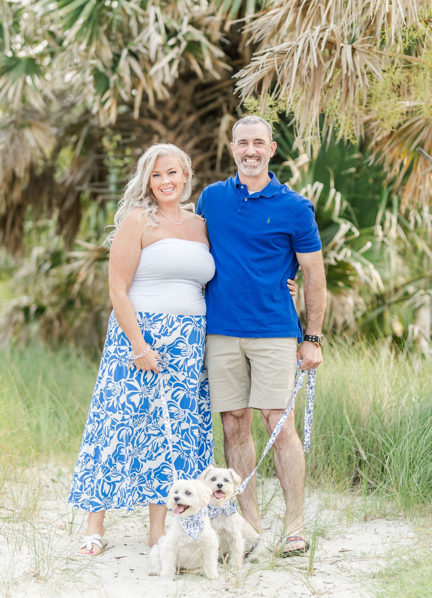 A couple poses with their small white dog on a beach path surrounded by palm trees.