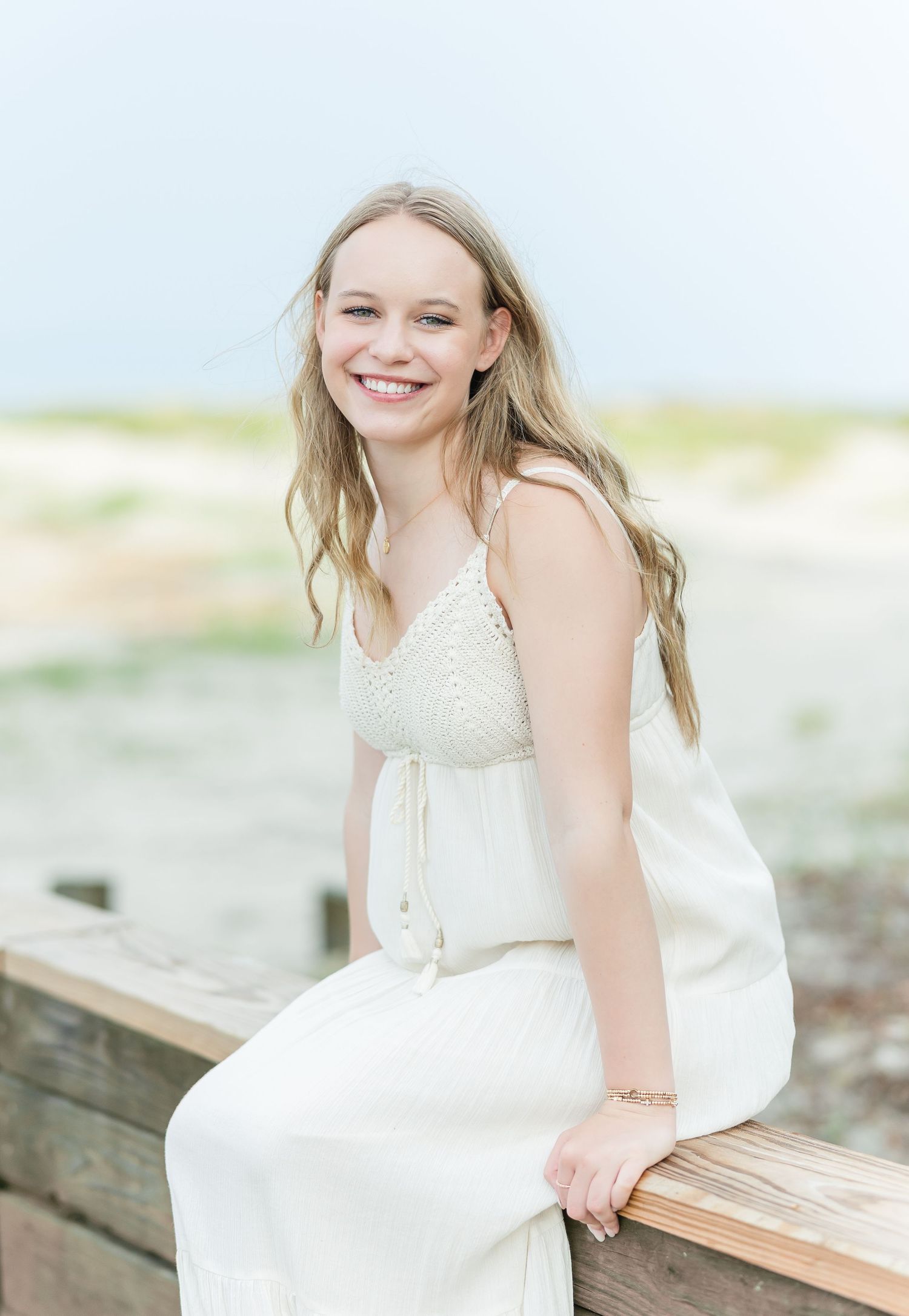 A person in a white summer dress sits on a wooden railing at the beach with wind-swept hair and a bright smile.