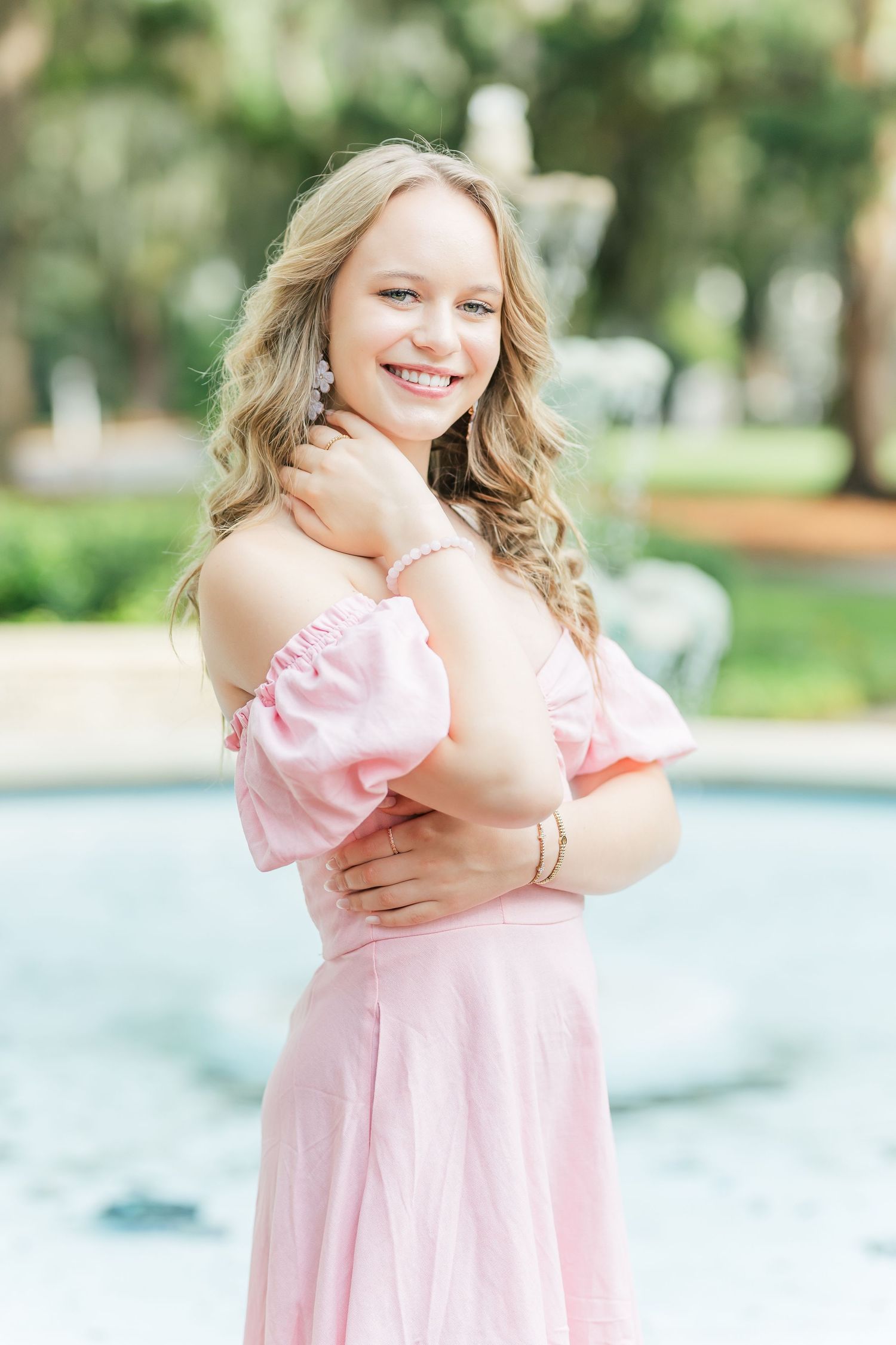 A person in a pink off-shoulder dress poses outdoors with a gentle smile in a garden setting.