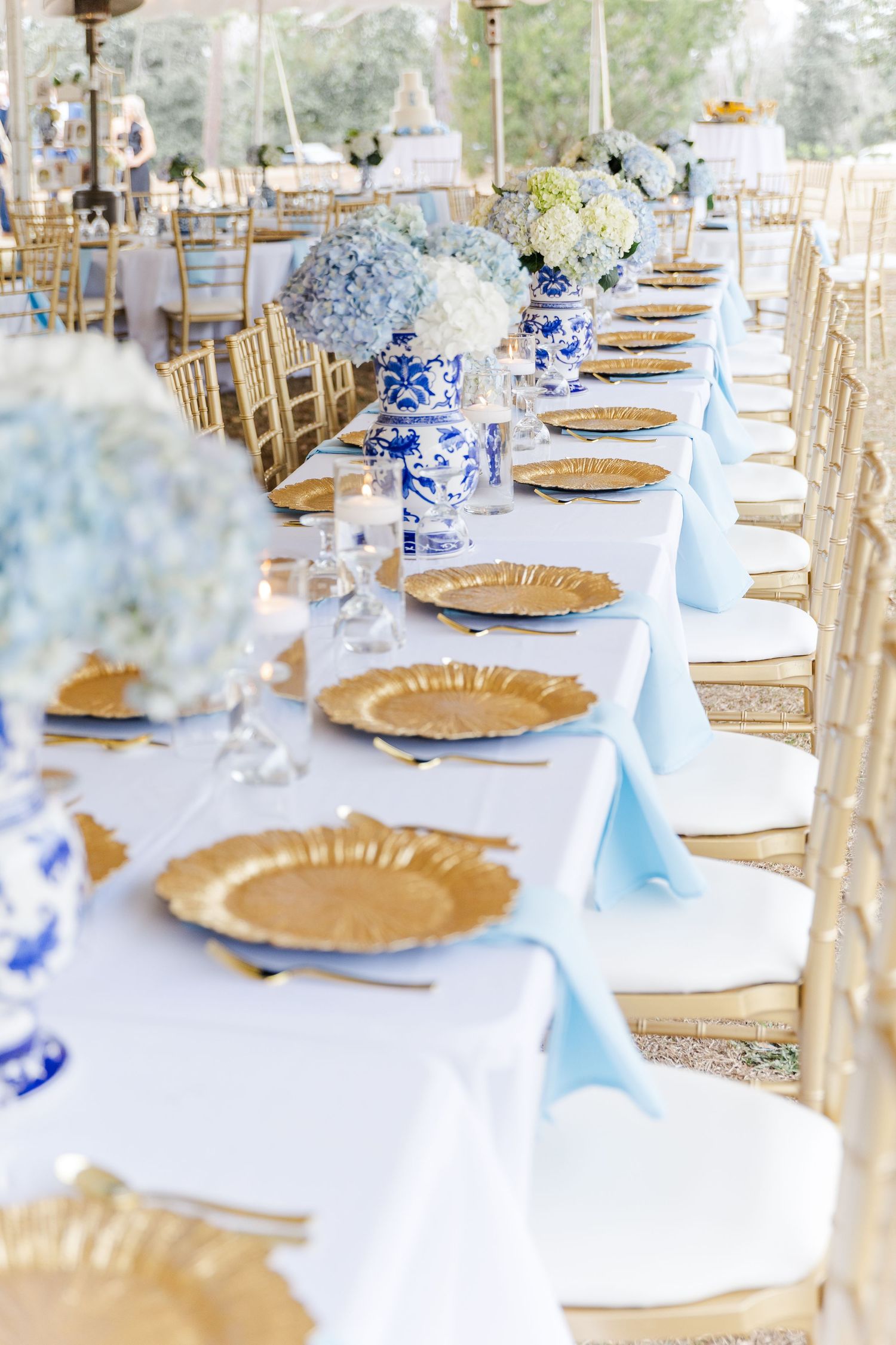 Elegant wedding reception table with gold charger plates, light blue napkins, and blue and white chinoiserie floral centerpieces.