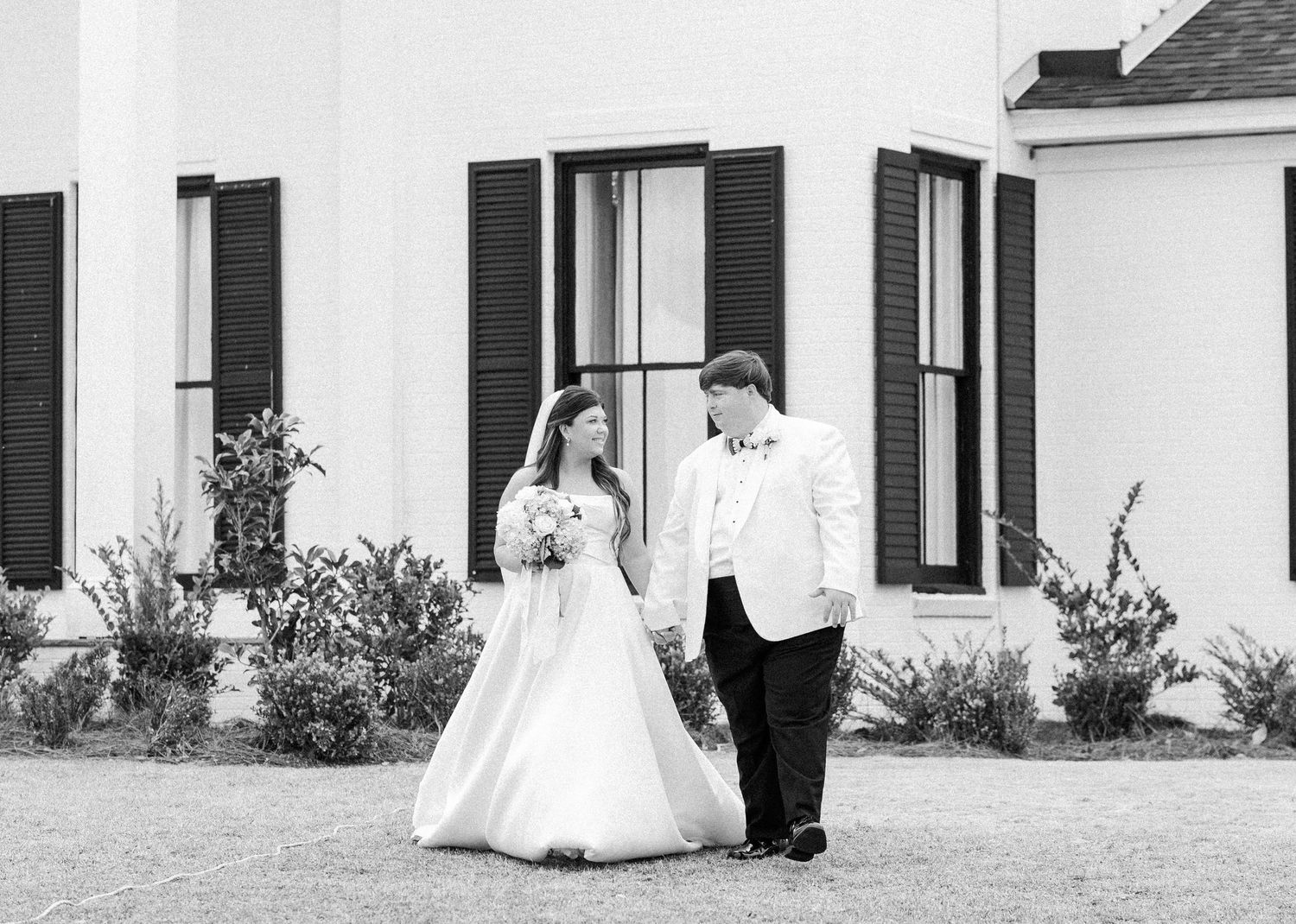 Bride and groom walk hand in hand in black and white photo outside white plantation-style venue with black shutters.