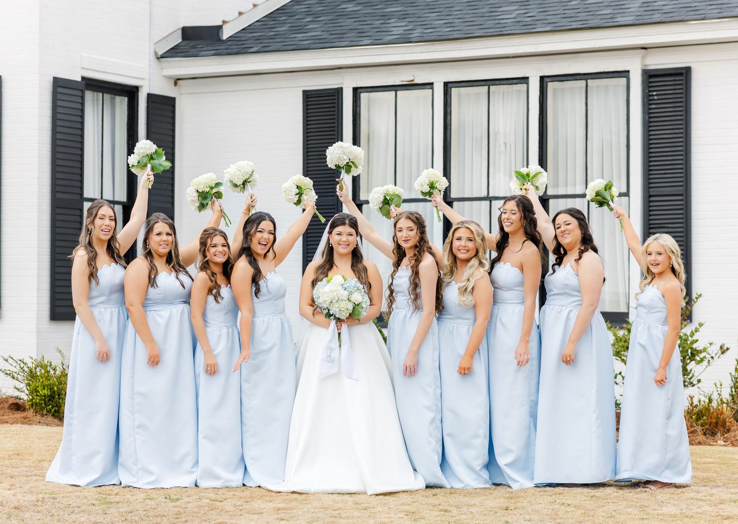 Bride with bridesmaids in light blue gowns celebrating outdoors raising white bouquets joyfully.