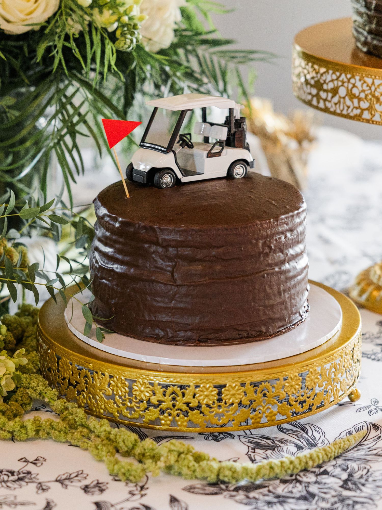 Chocolate groom's cake with golf cart topper on gold cake stand with greenery decor.