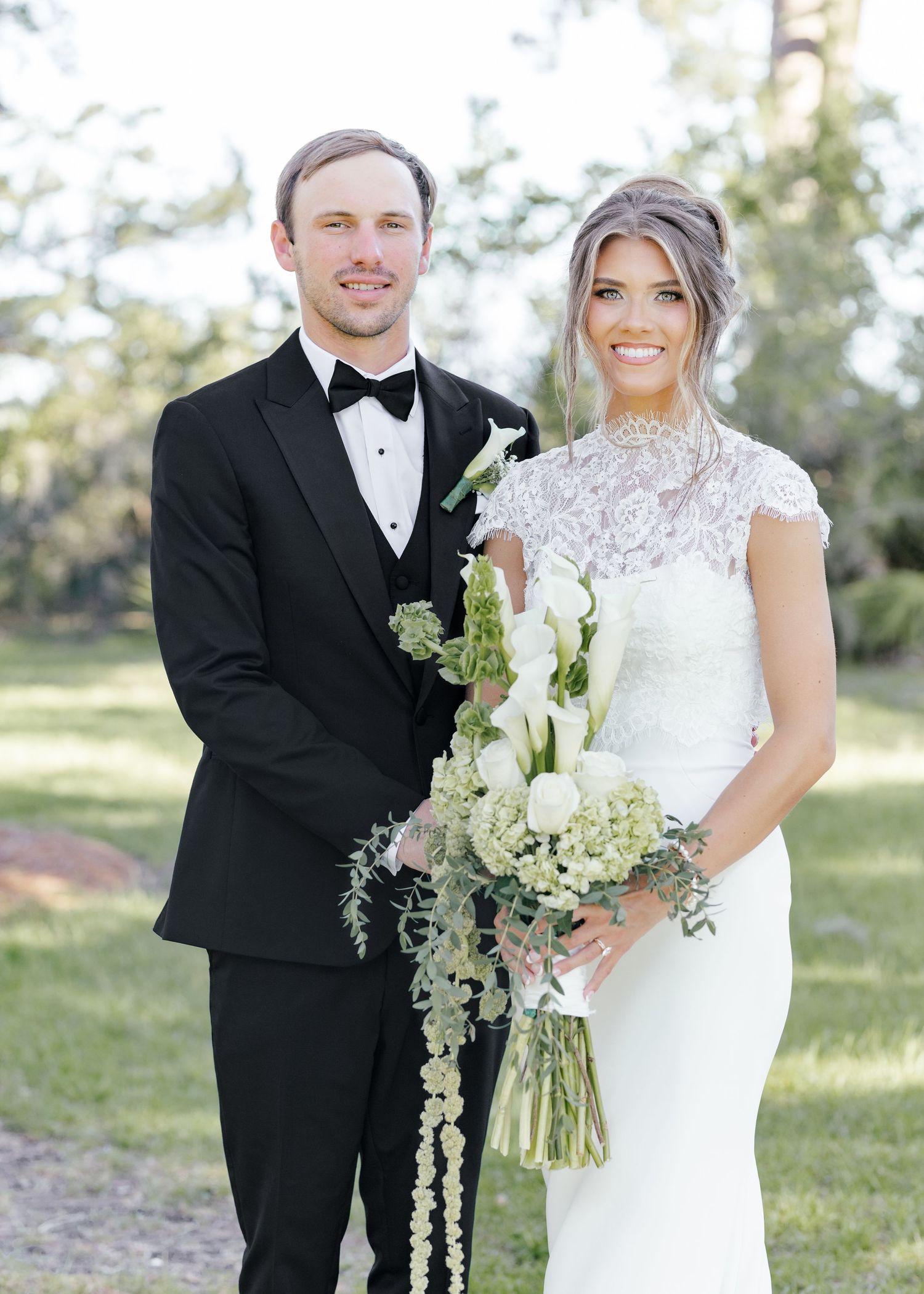Wedding couple posing outdoors, groom in black tuxedo, bride in lace gown holding white floral bouquet.