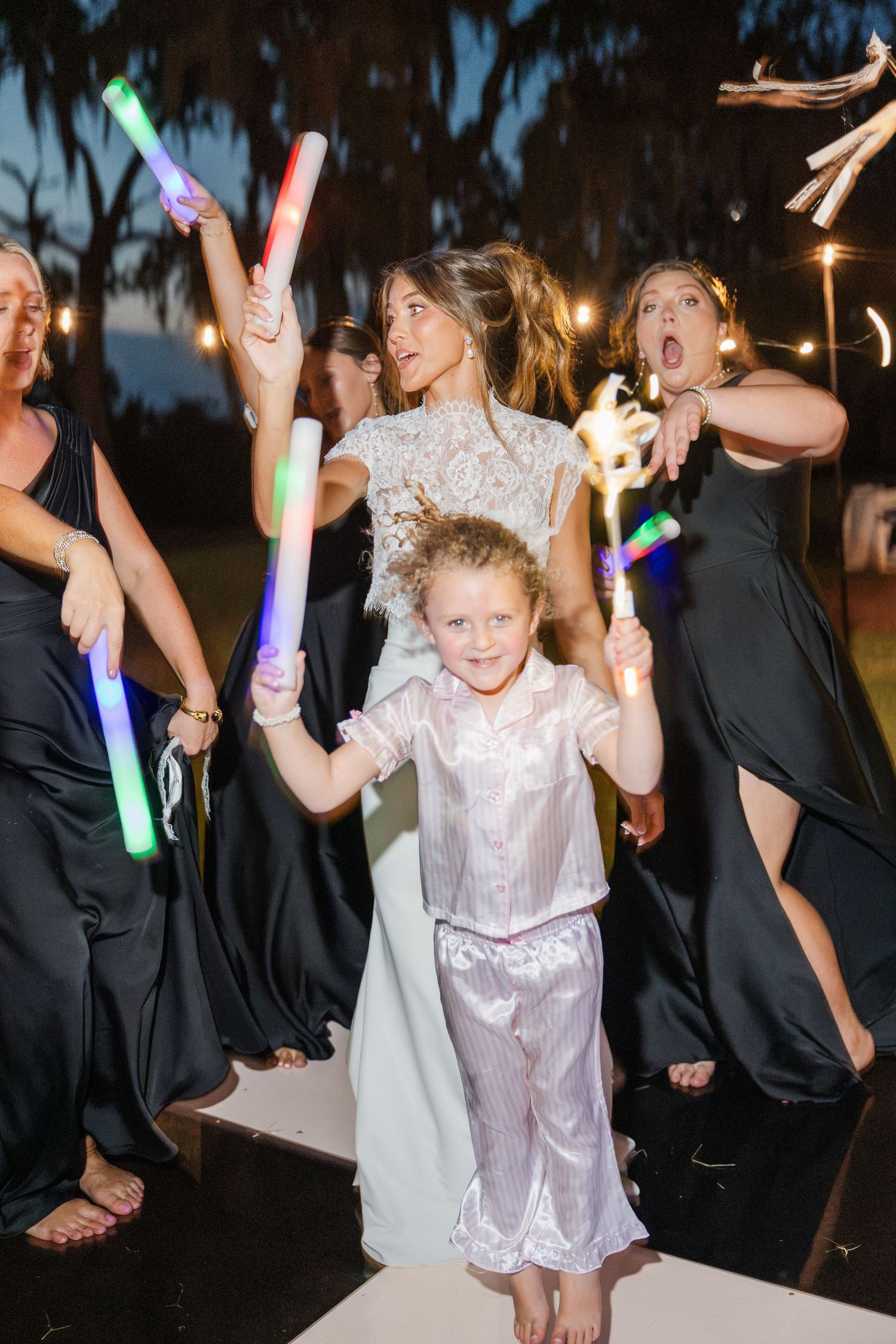 Bride in lace gown holds glowing foam stick with flower girl and bridesmaids in black dresses at outdoor wedding reception.