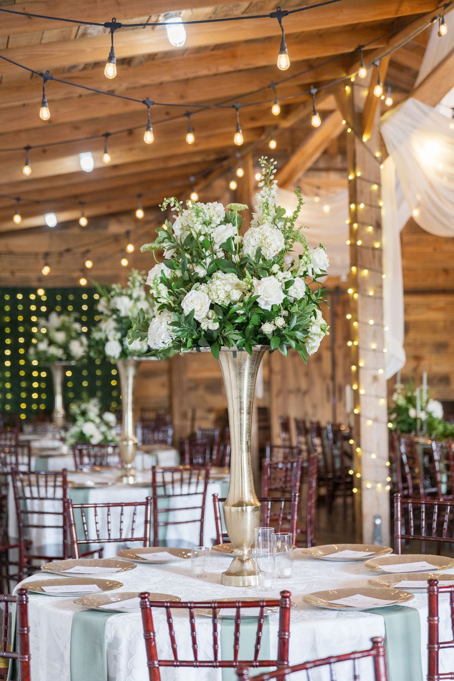 Elegant barn wedding reception setup with white floral centerpieces, string lights and mahogany chiavari chairs.