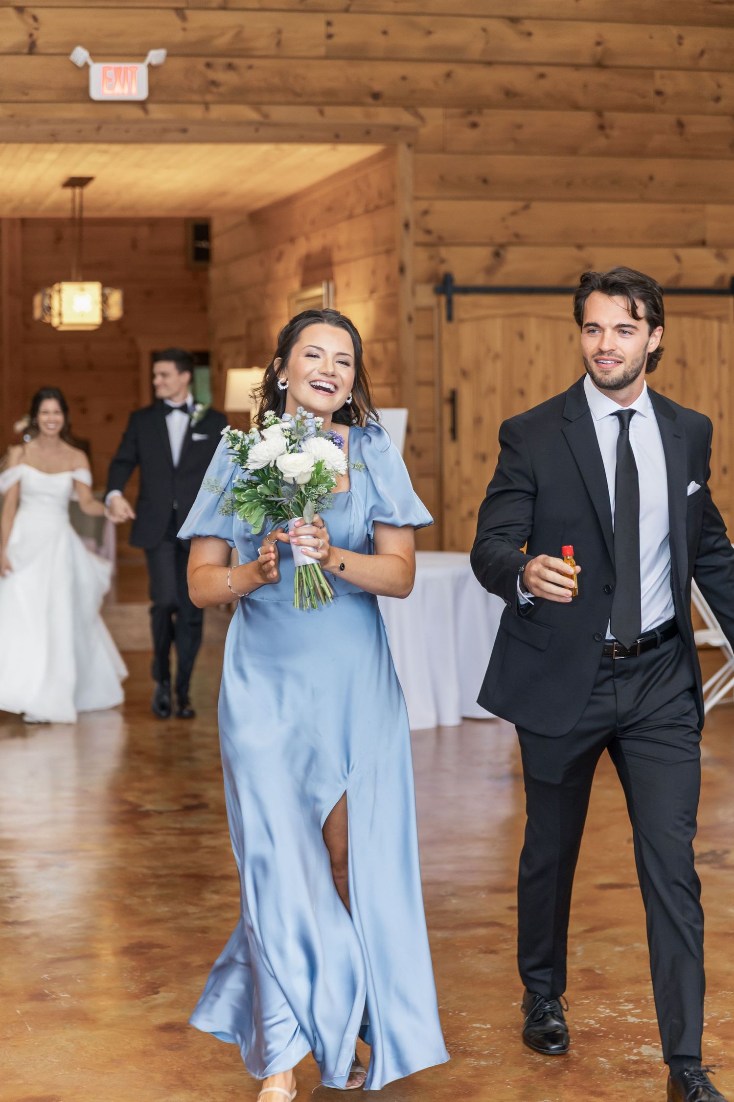 Wedding party members in light blue dresses and black suits walk across a rustic wooden venue floor.