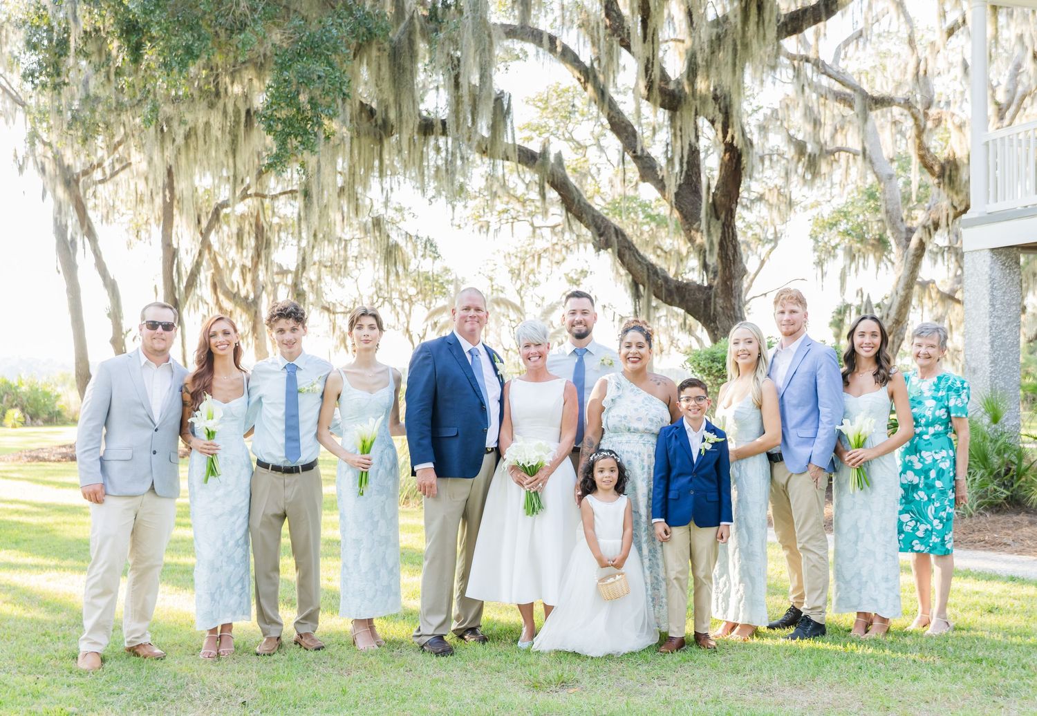Wedding party in light blue and white attire poses together under Spanish moss-draped trees at outdoor venue.