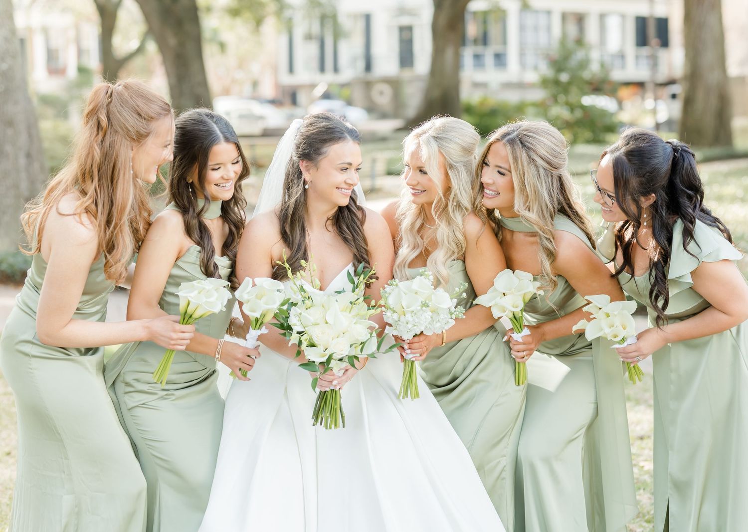 Bridesmaids in soft sage green dresses stand together in a line holding white bouquets.