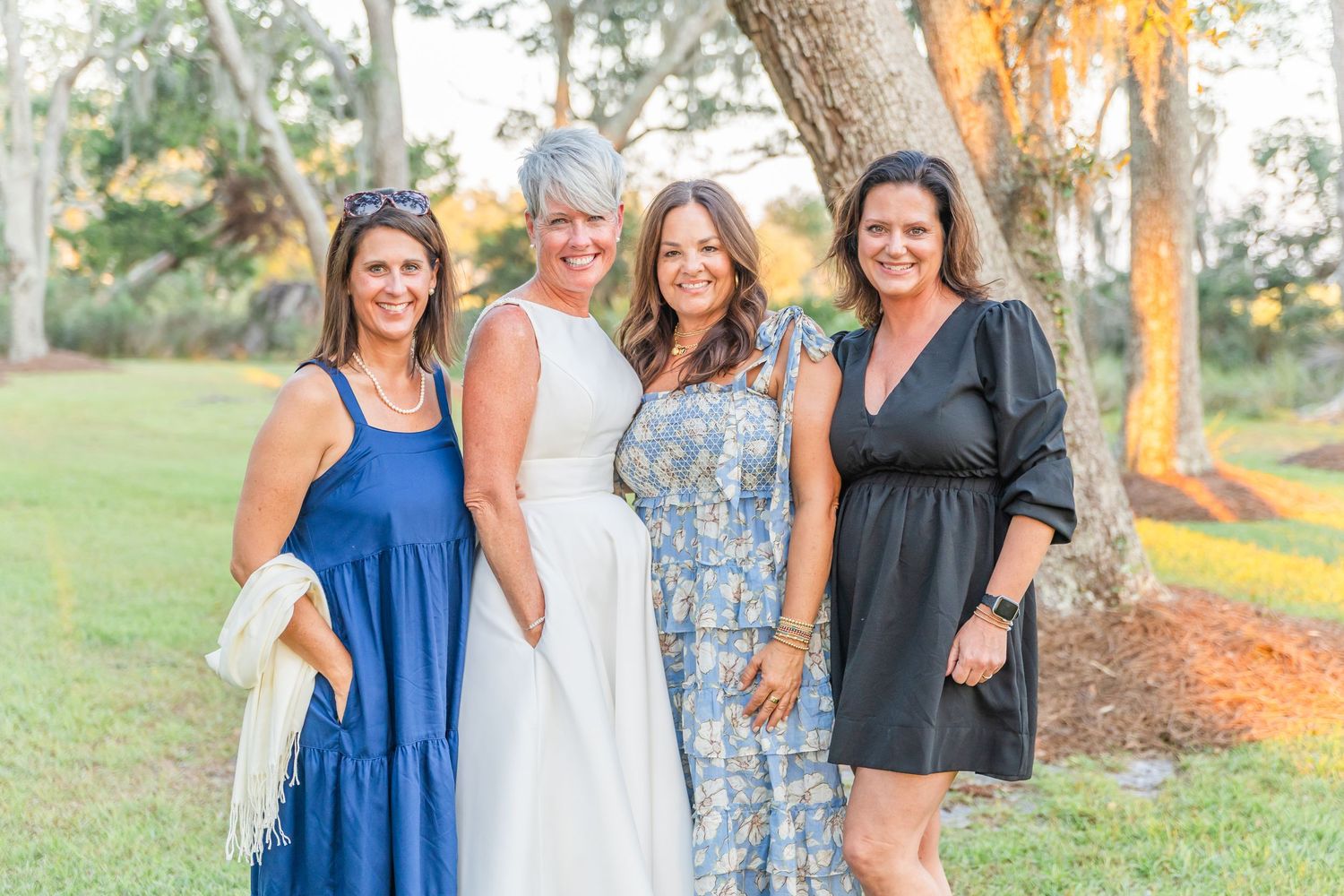 Four women pose together outdoors at a summer event wearing colorful dresses.