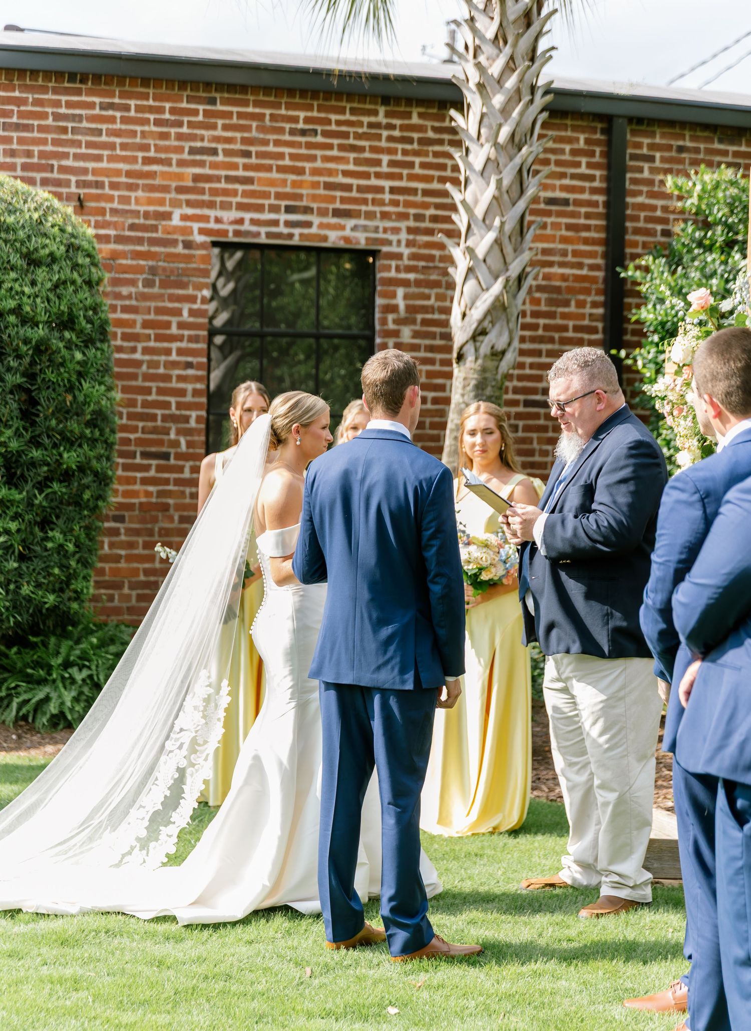 Wedding party gathers near a brick building with palm trees during an outdoor celebration.