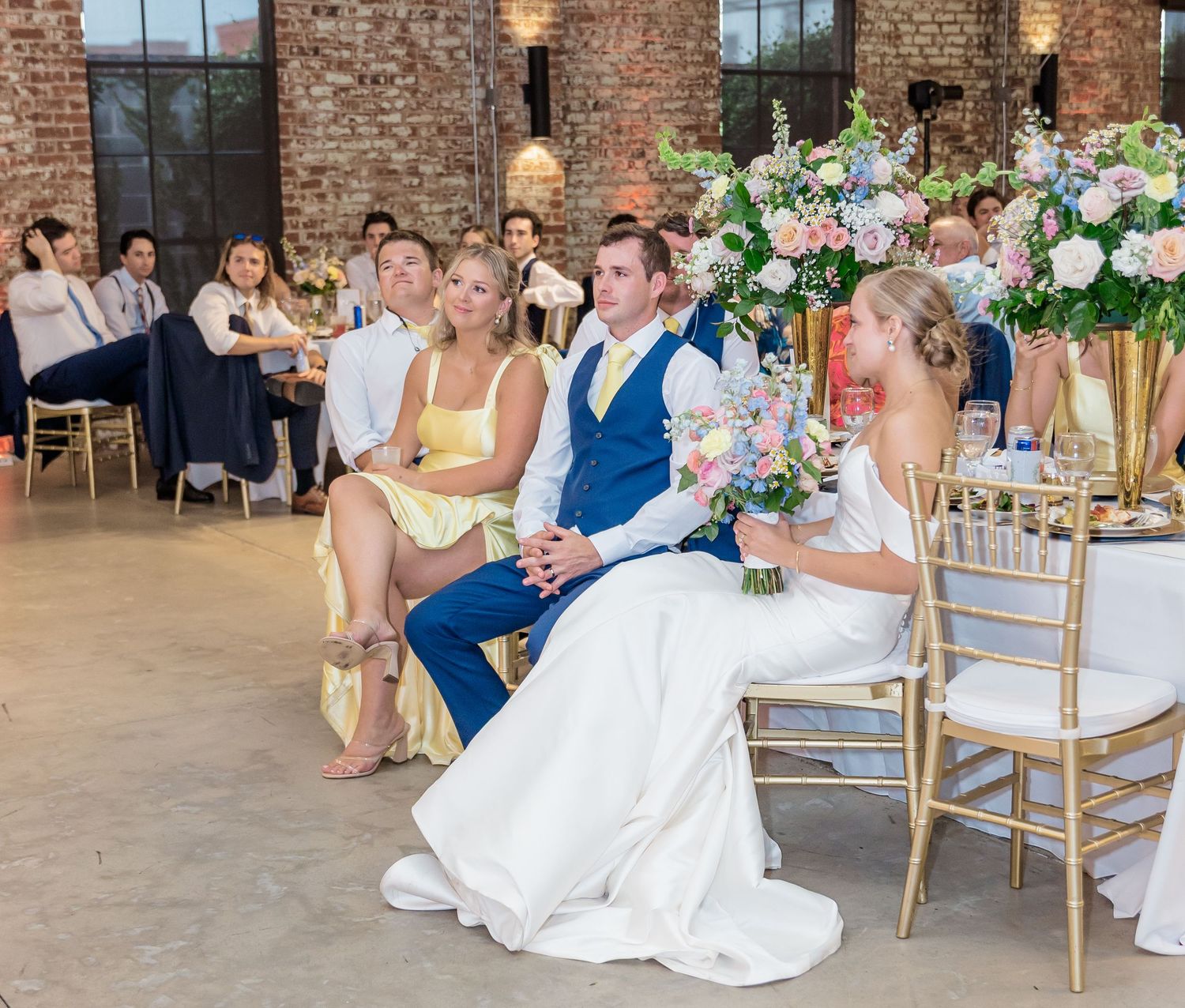 Wedding reception setup with white floral centerpieces in an elegant brick-walled venue with gold chairs.