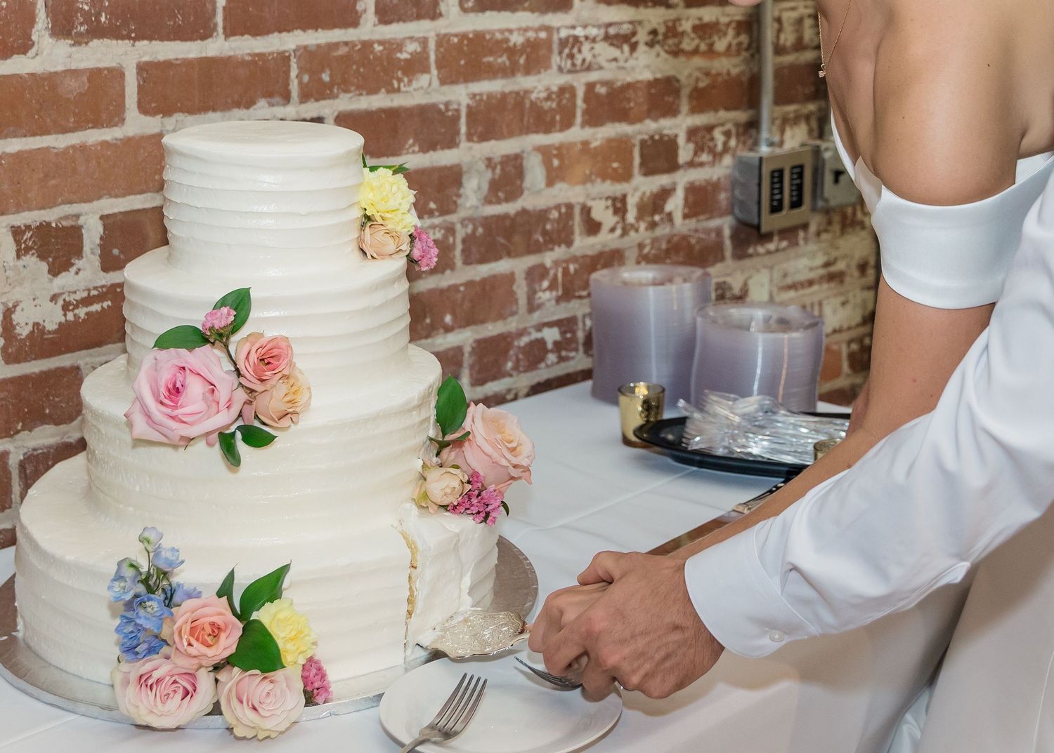 Hands cutting into a white wedding cake decorated with pink and blue flowers against a rustic brick wall.