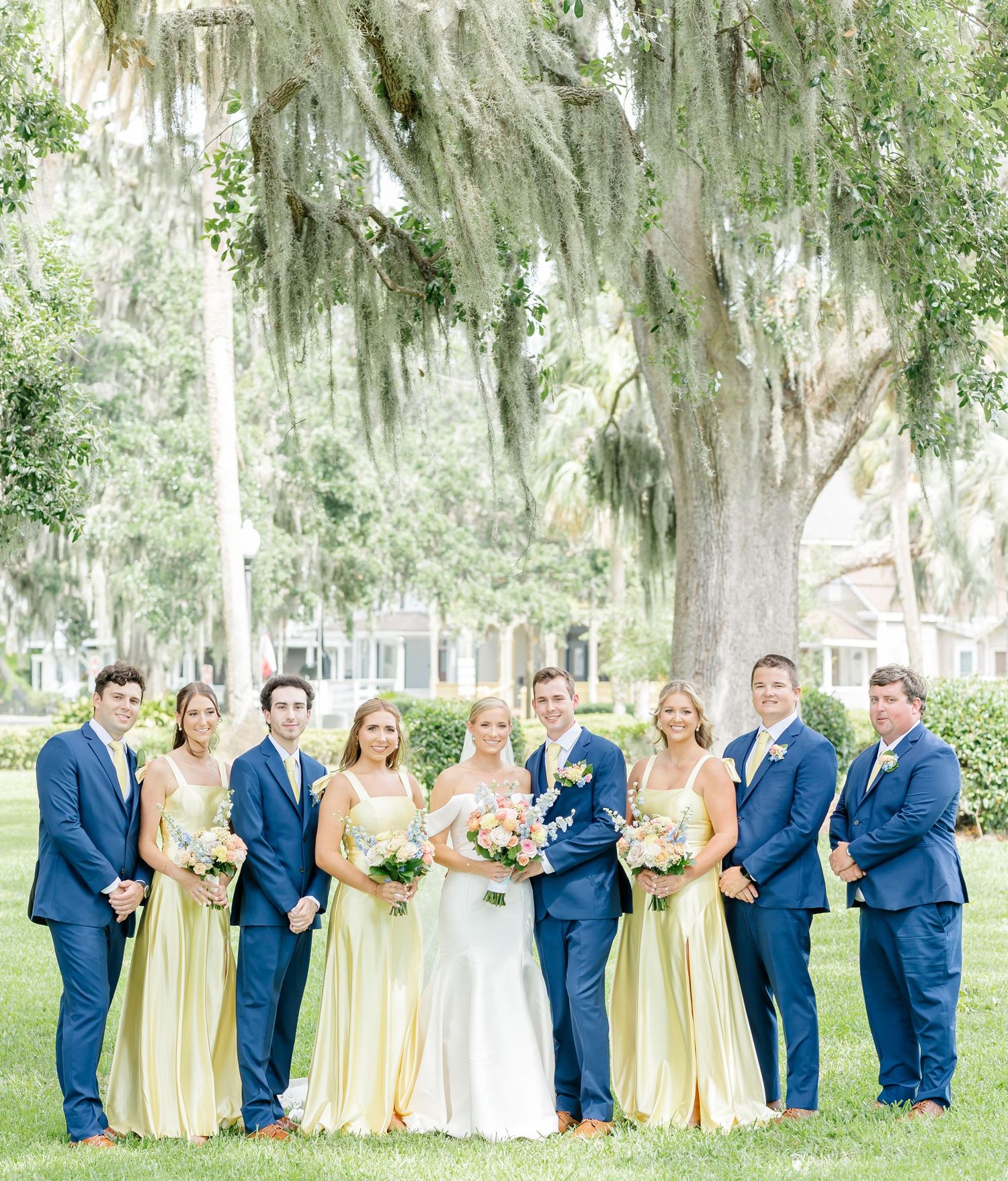 Wedding party in navy blue suits and pale yellow dresses pose together on a lawn with Spanish moss trees in background.