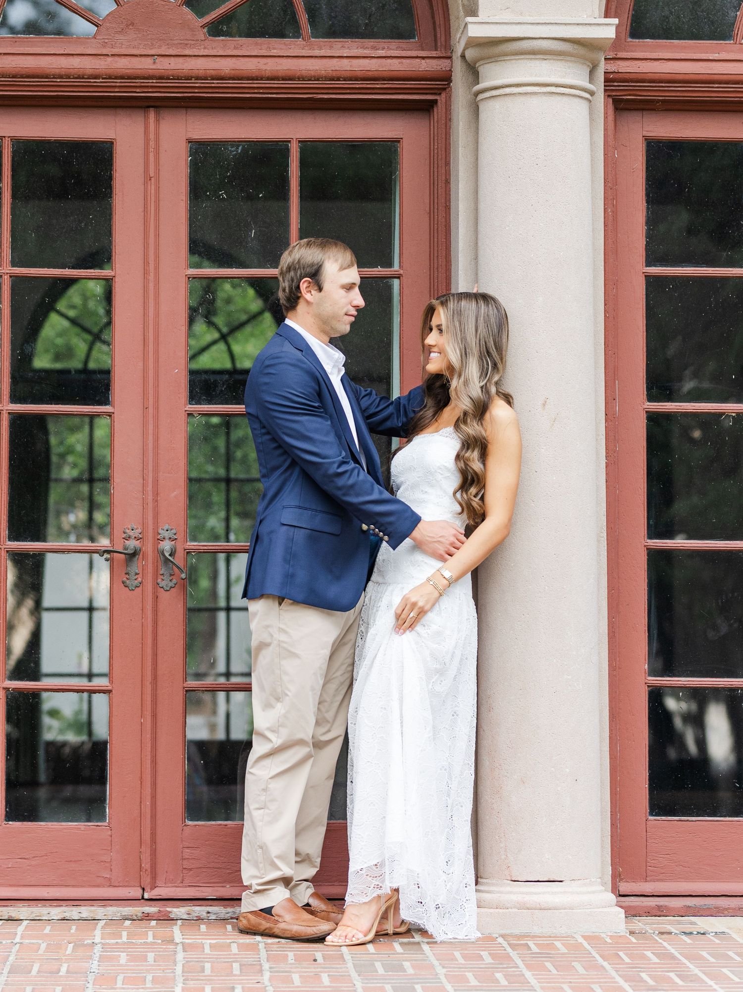 Couple in engagement photos, man in navy blazer and woman in white lace dress pose by elegant column.