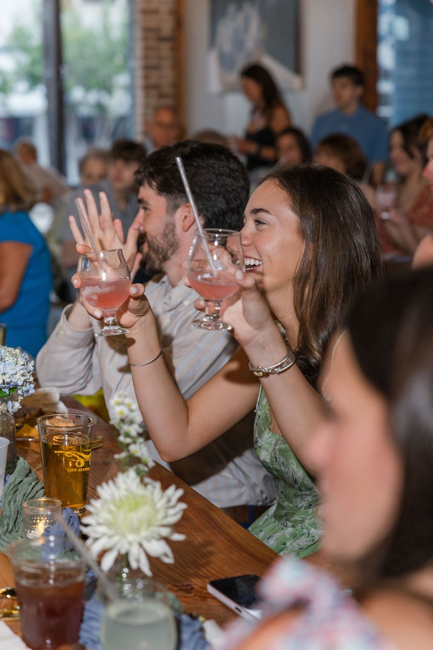Couple laughing and holding pink cocktails at a lively restaurant gathering with guests in the background.