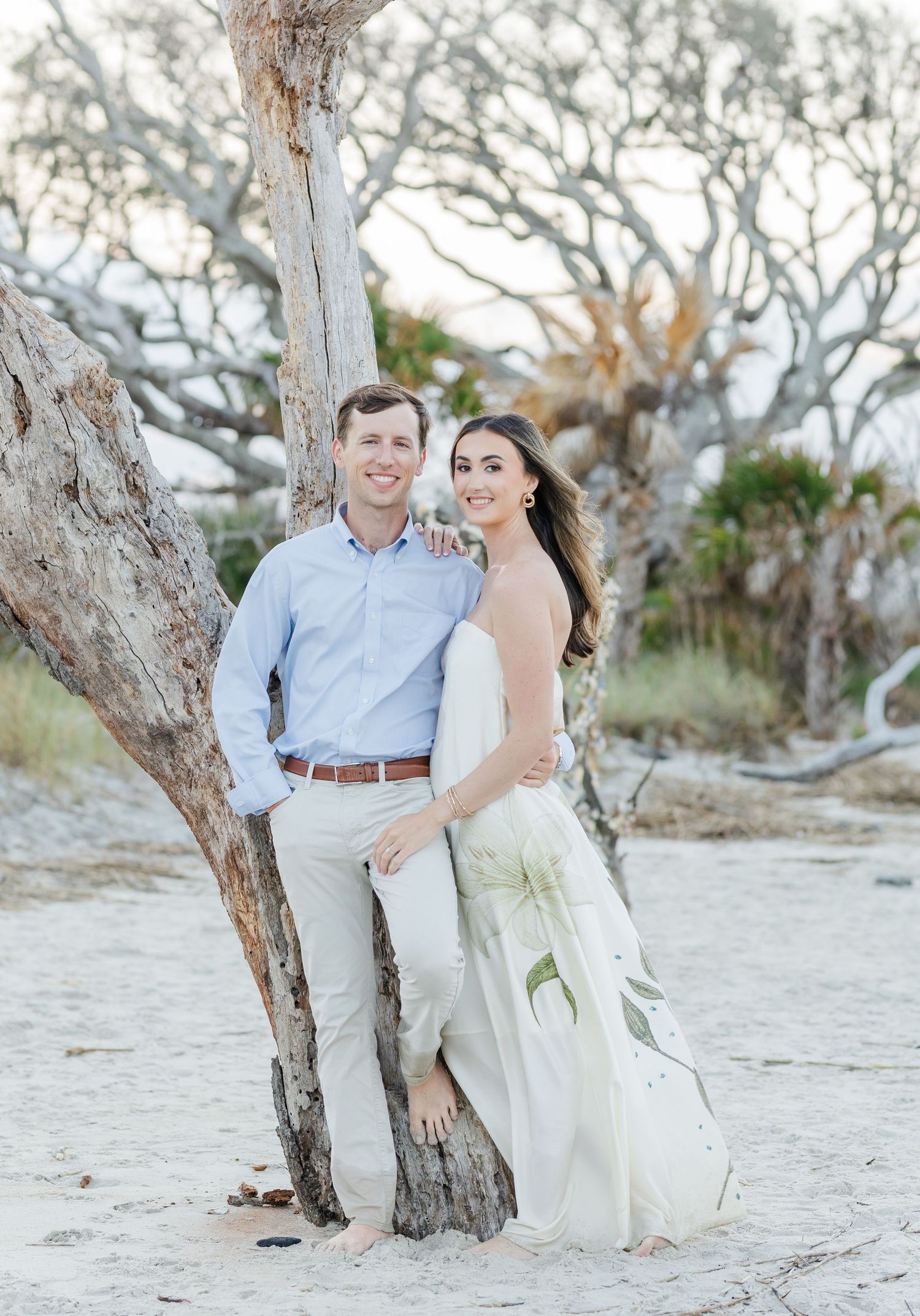 Smiling couple posing together against a weathered driftwood tree on a sandy beach during engagement photos.