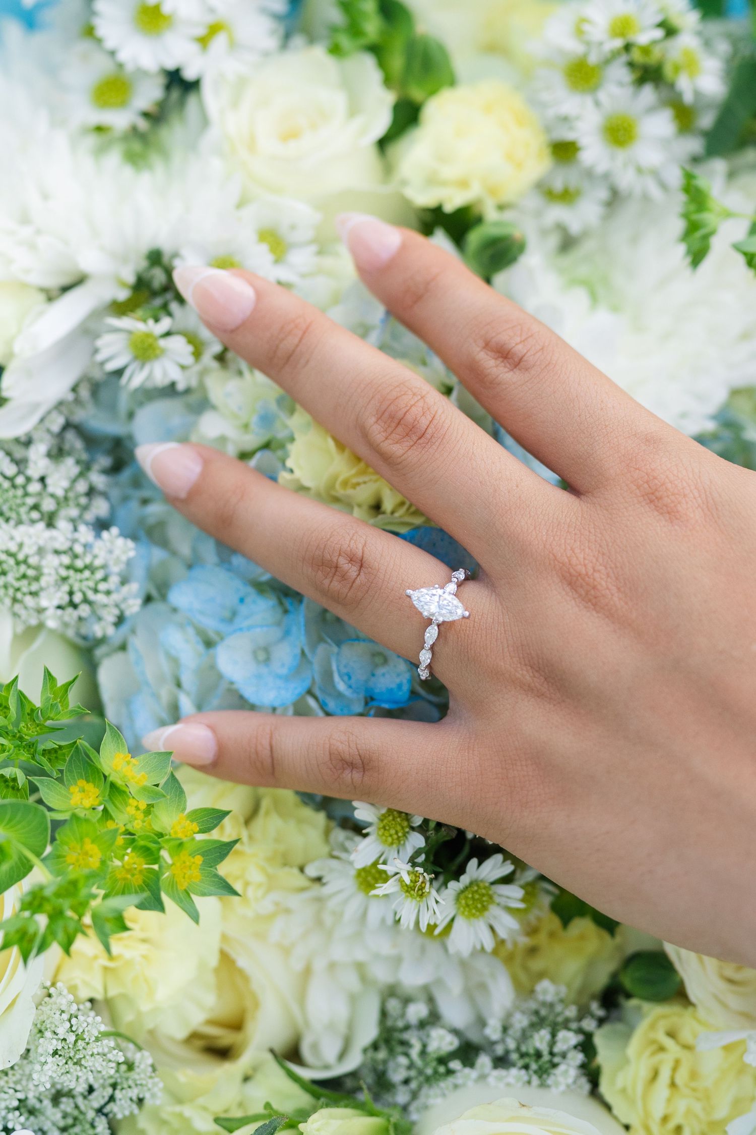 Close-up of a hand displaying a marquise diamond engagement ring resting on a colorful floral bouquet.