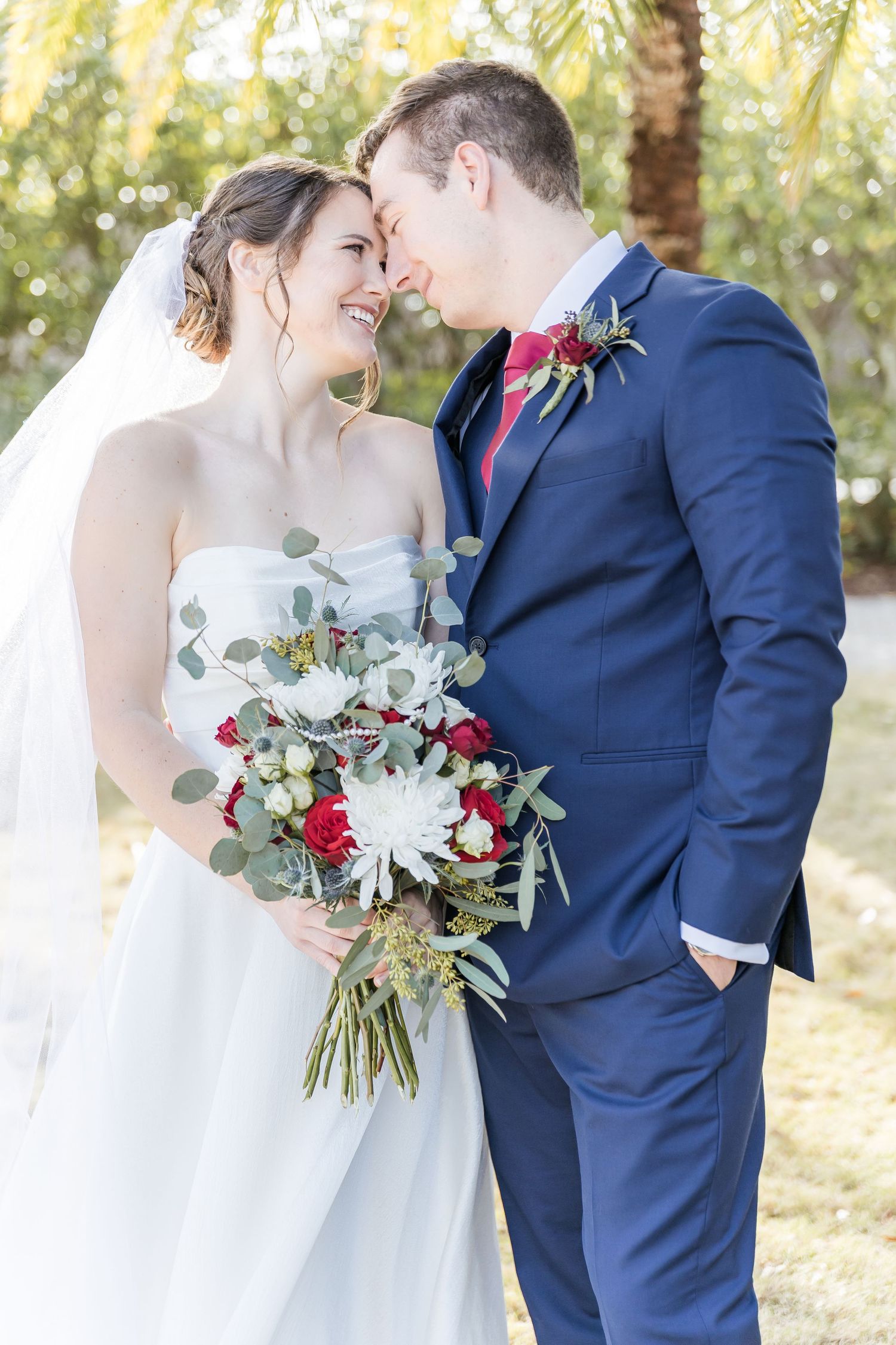 Romantic wedding couple embraces outdoors with bride holding white and red bouquet against palm trees.