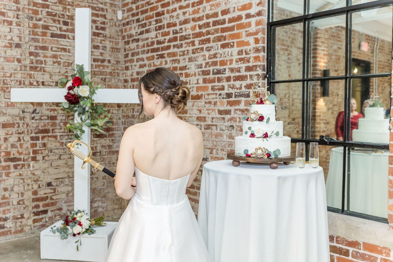 A wedding dress and multi-tiered white cake in a rustic brick venue with a floral-decorated cross.