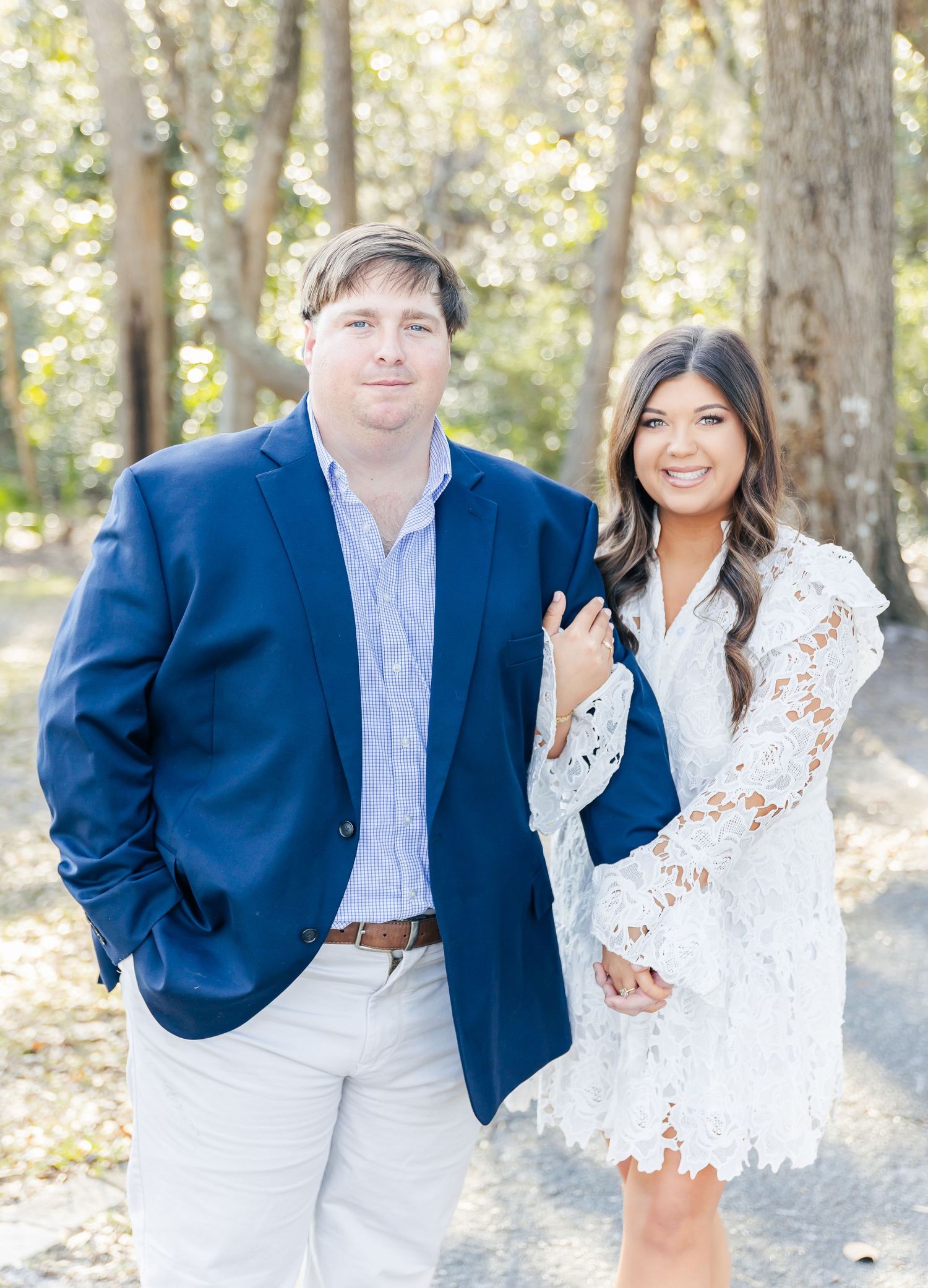 Engagement photo featuring a couple in coordinating navy and white outfits on a sun-dappled forest path.