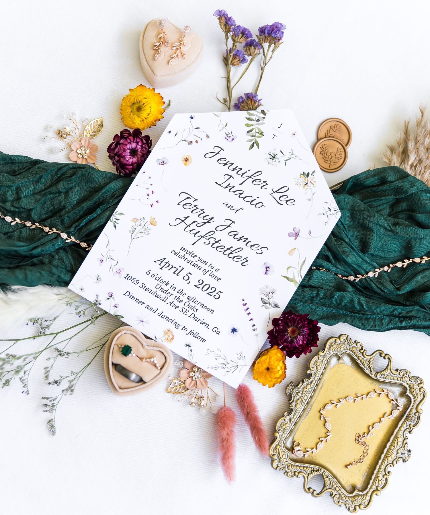 Wedding invitation with floral design displayed with dried flowers, antique frames, and emerald green fabric.