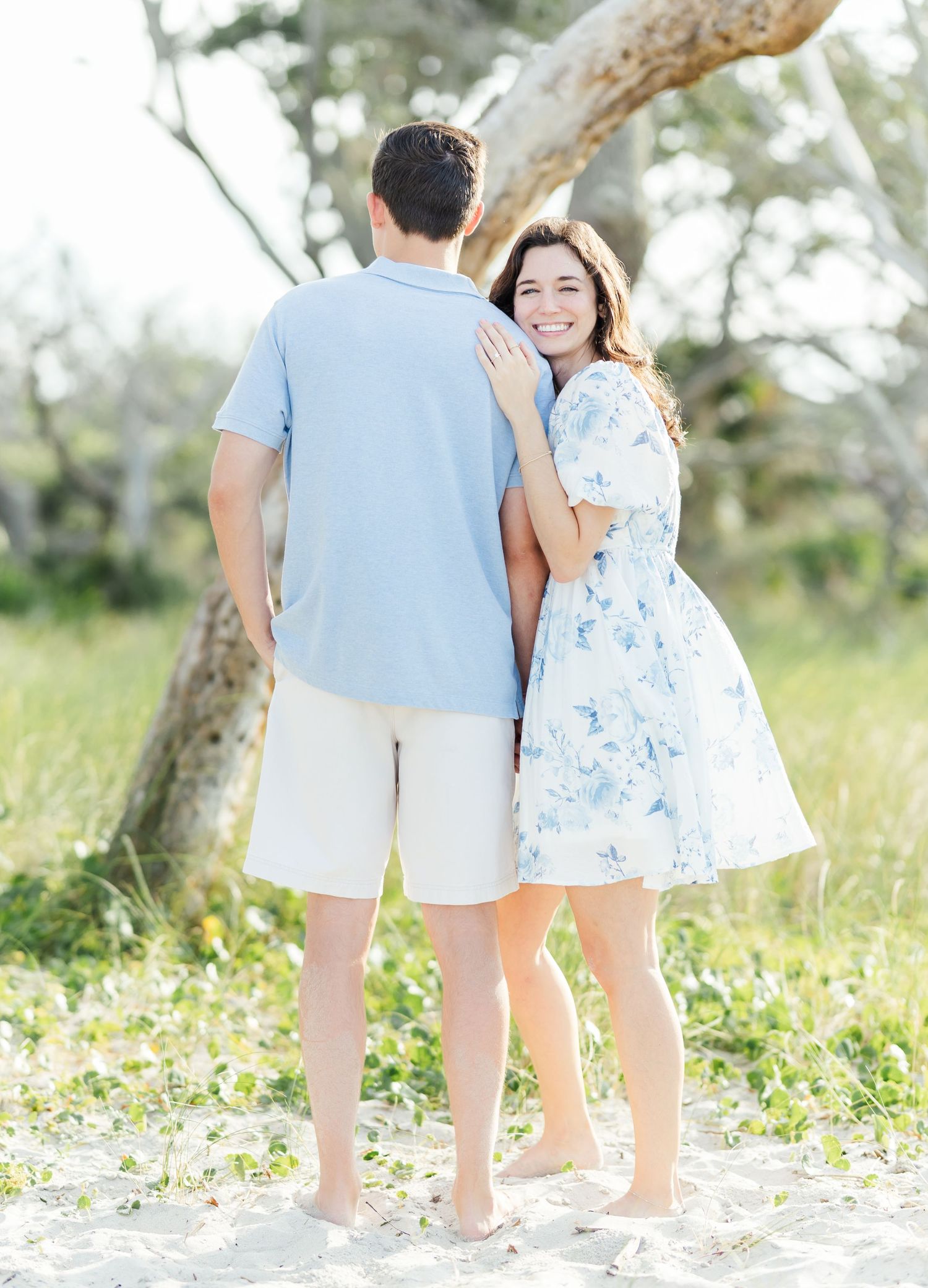 Romantic engagement photo of couple in blue and white outfits standing barefoot near a tree.
