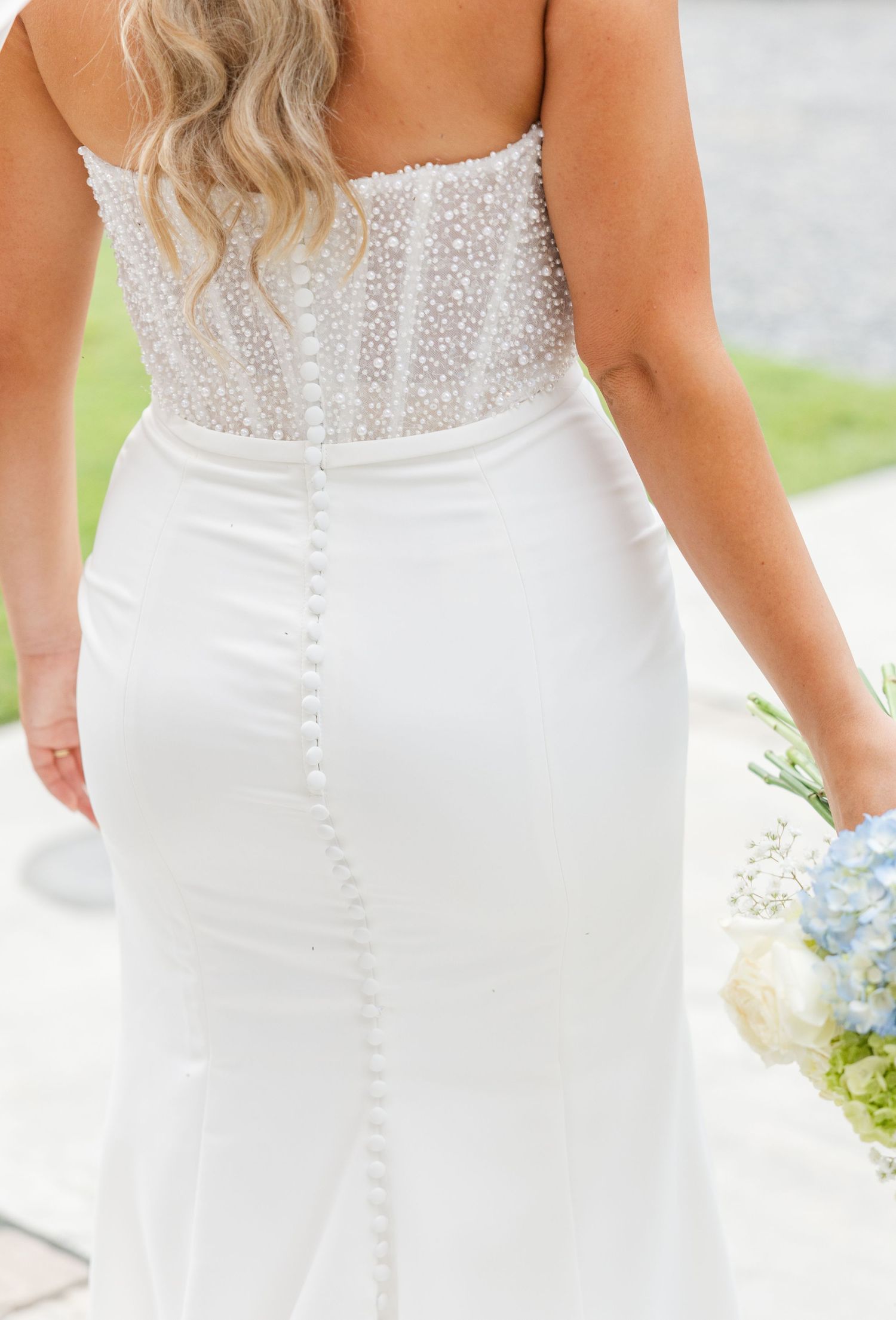 Back view of bride in strapless pearl-embellished wedding gown with button detail, holding blue and white bouquet.