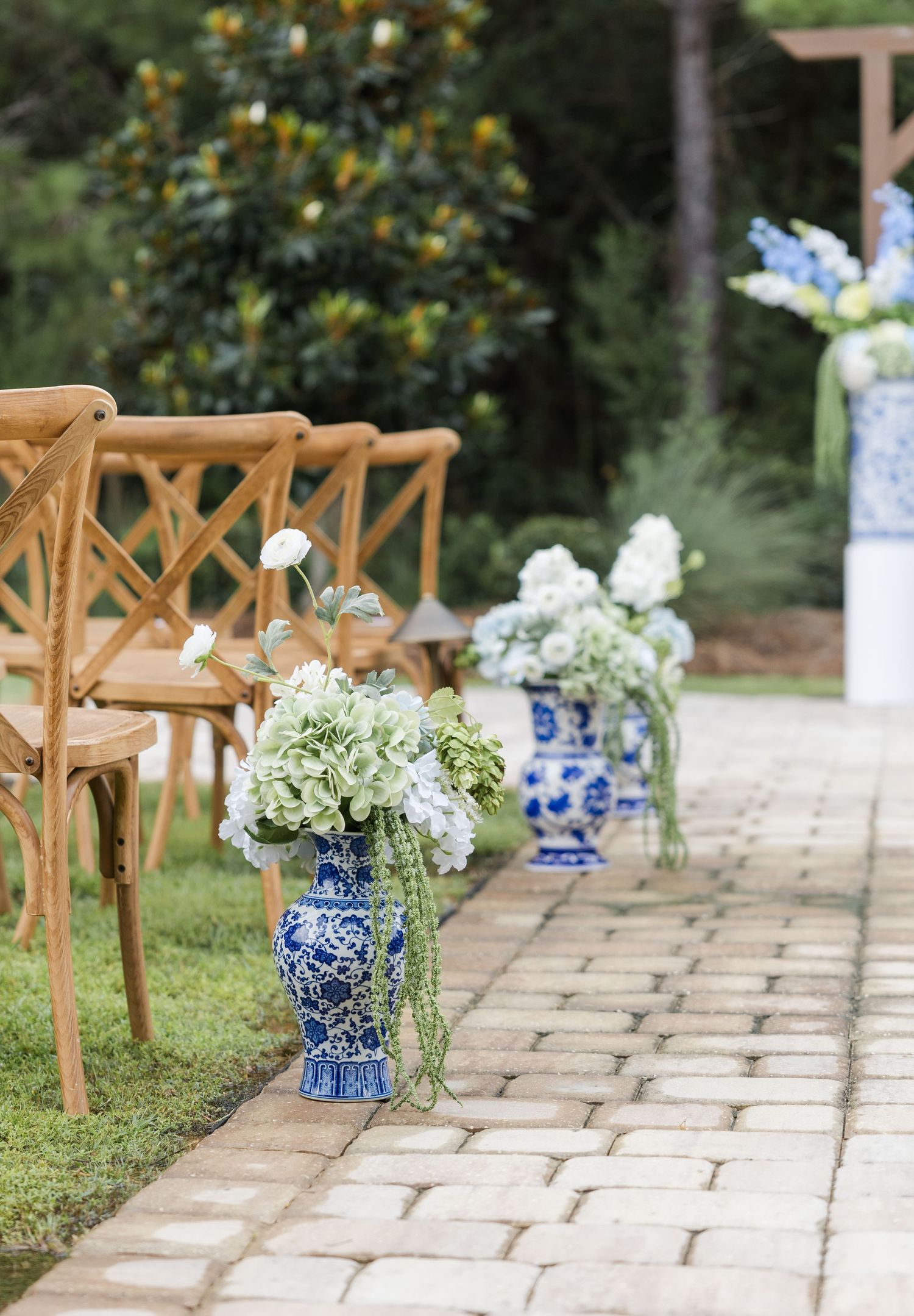 Blue and white chinoiserie vases with white hydrangeas lining a brick wedding ceremony aisle with wooden chairs.