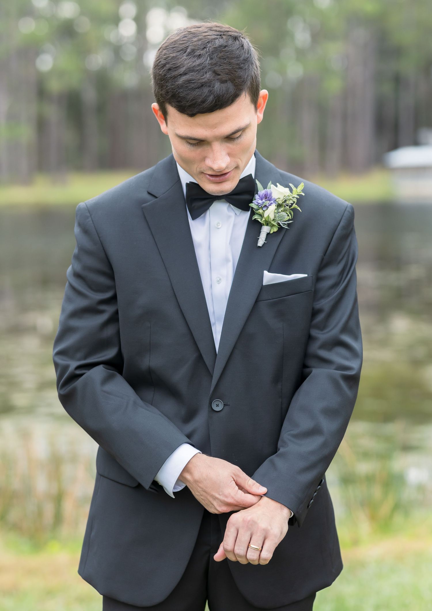 Close-up detail shots of a black tuxedo with bowtie and boutonniere against a natural lakeside setting.