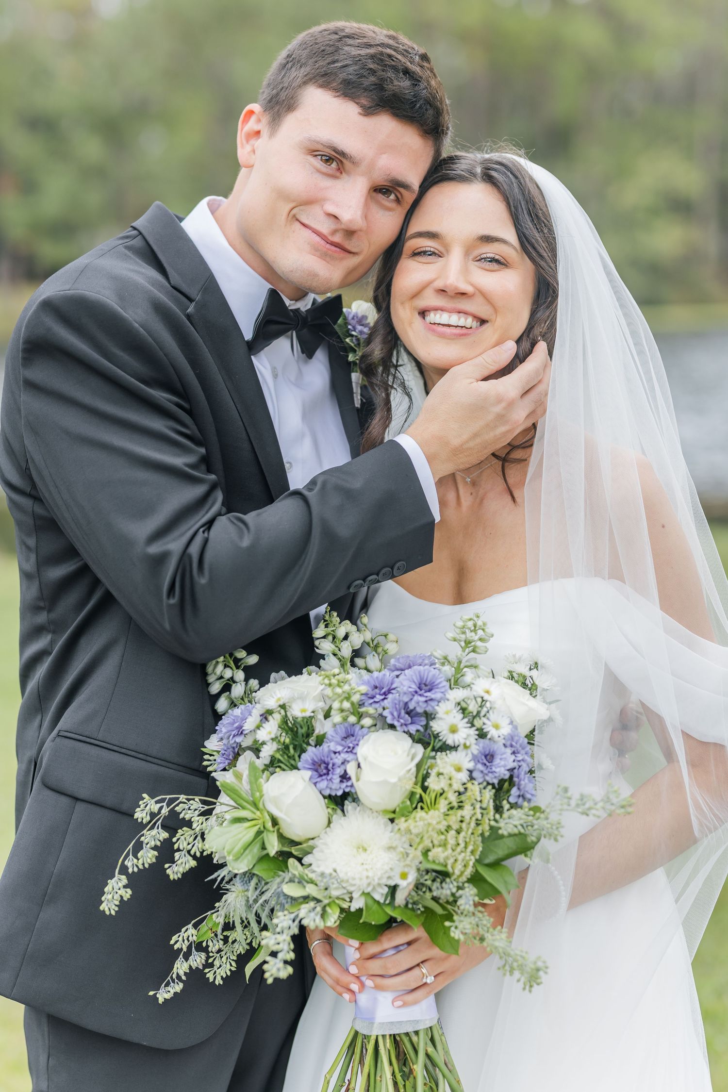 Newlyweds share tender moments while holding a purple and white bouquet in a garden setting.