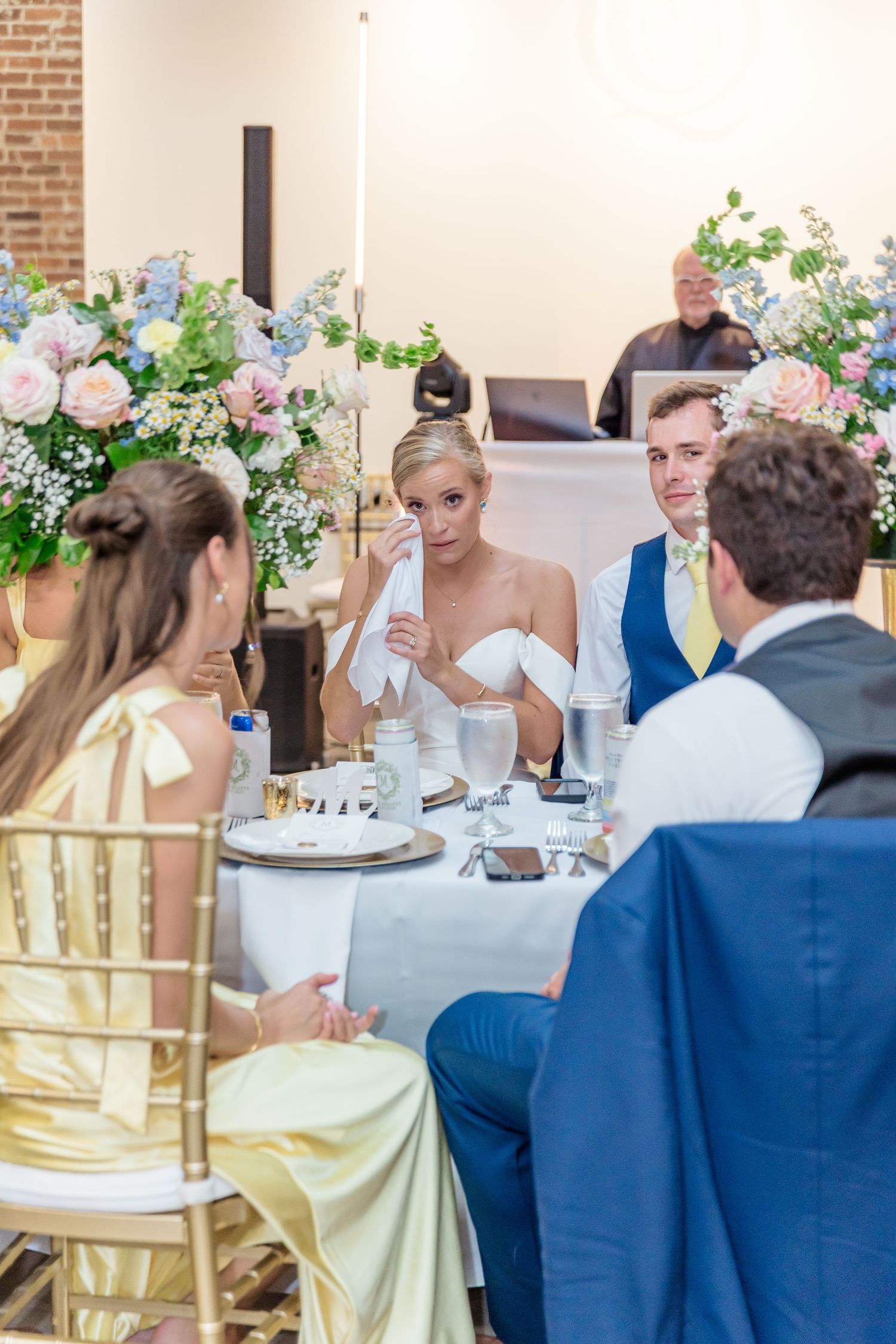 Wedding reception table setting with yellow dresses and blue suits at a rustic brick venue with floral centerpieces.