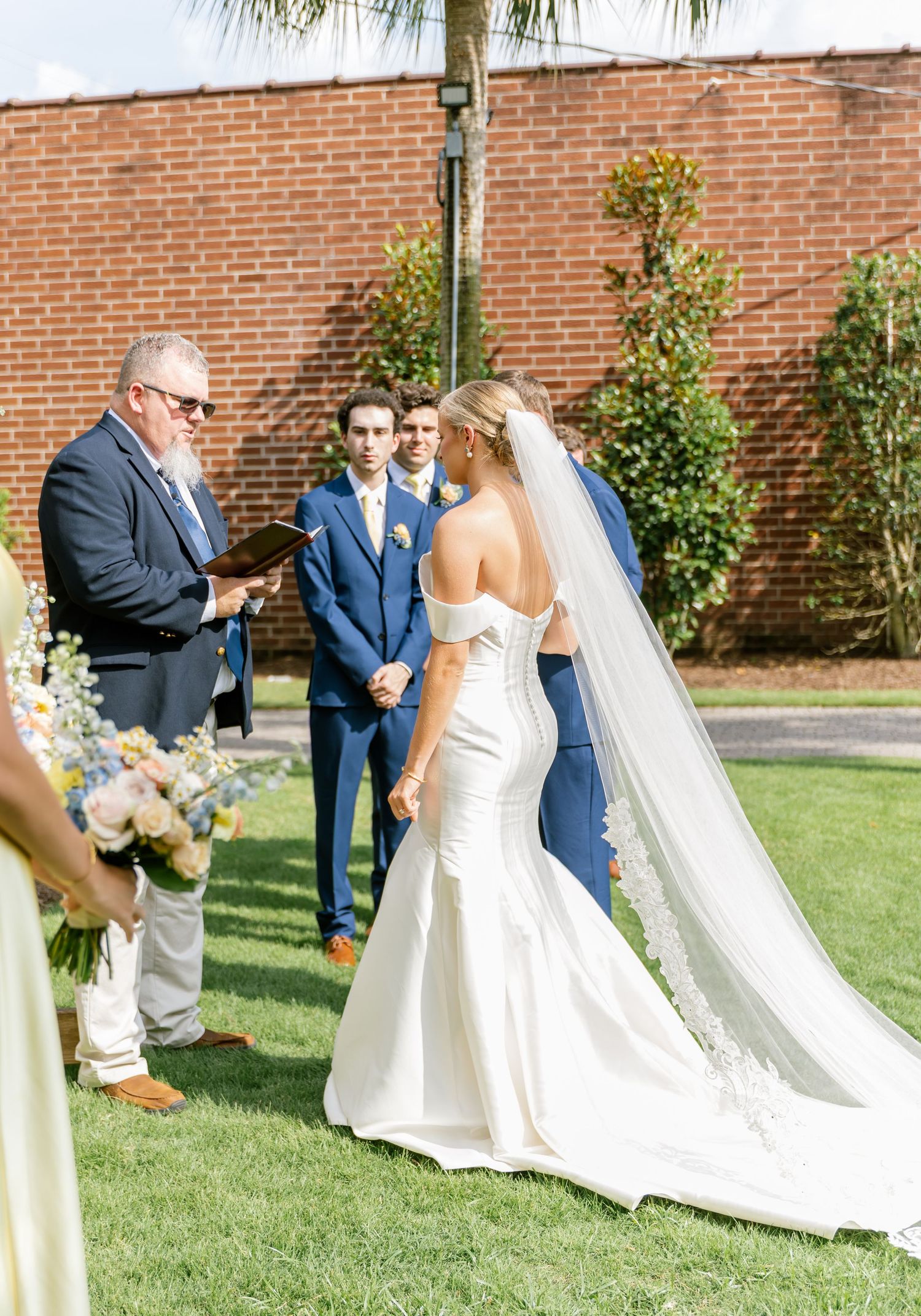 Wedding party members in formal attire line up on a green lawn against a brick wall backdrop during an outdoor ceremony.
