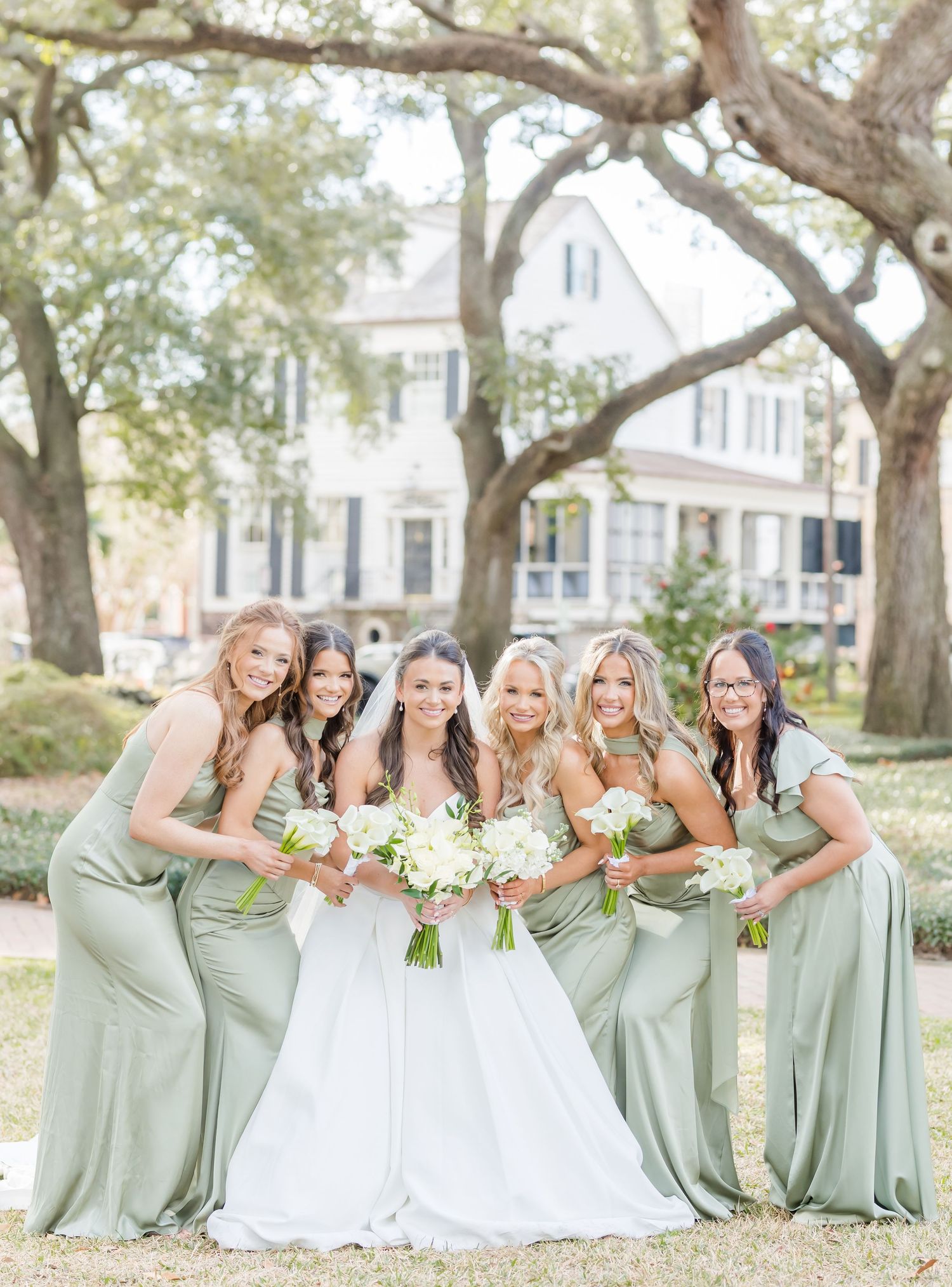 Bridesmaids in sage green dresses pose playfully with the bride in a white gown during an outdoor wedding photoshoot.