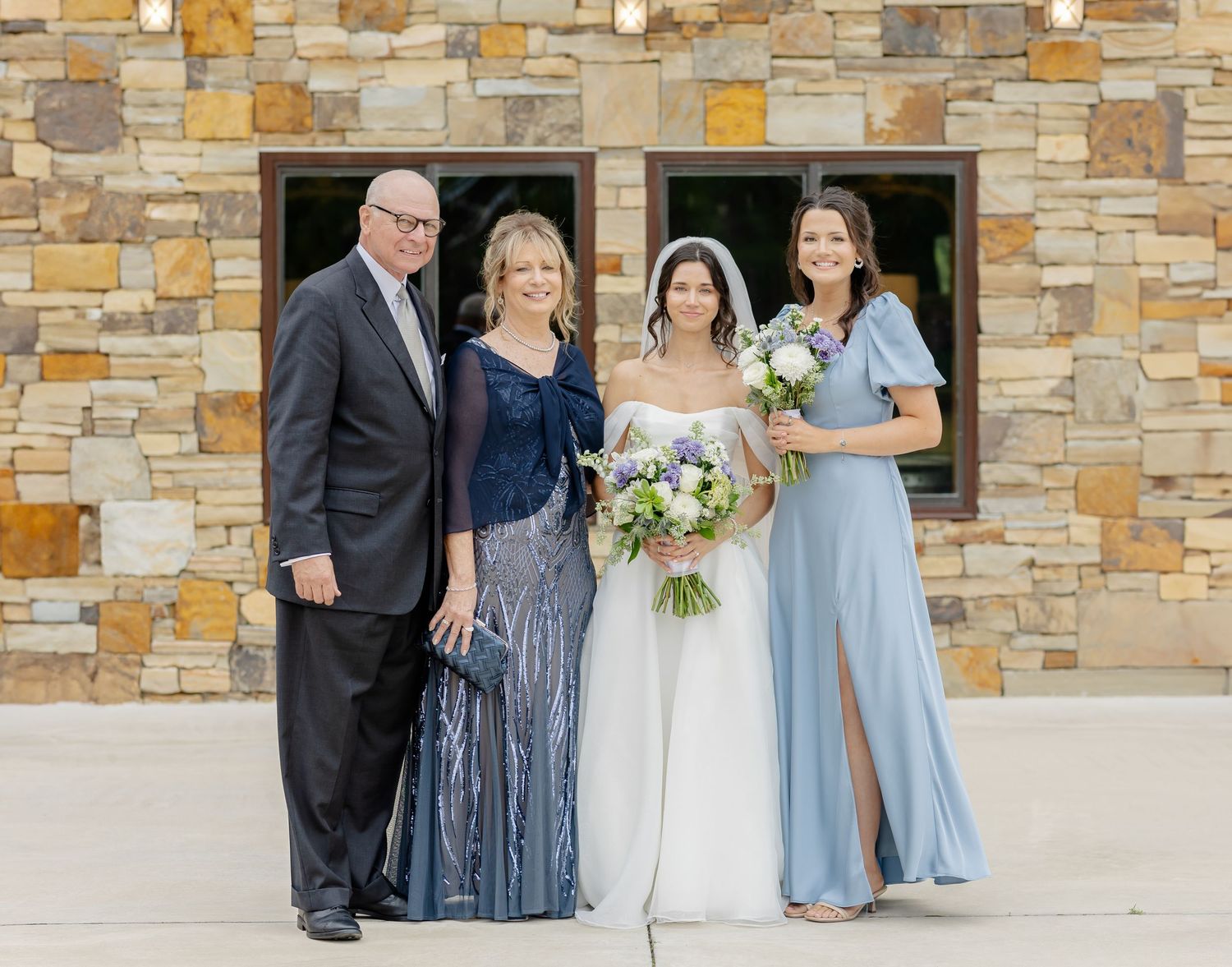 Wedding party members in formal attire pose together against a stone wall exterior.