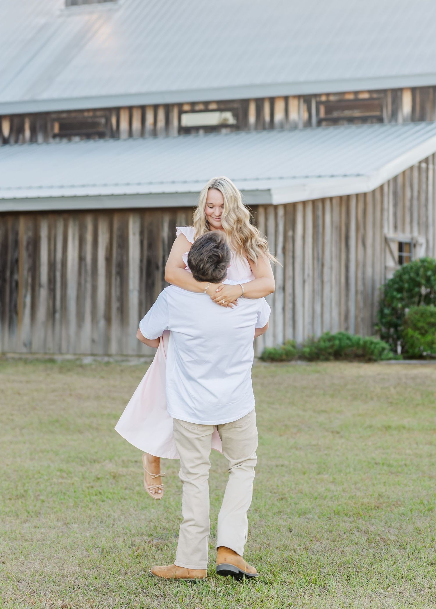 Two people in white shirts and khaki pants dance playfully together on a grassy lawn near a wooden fence.