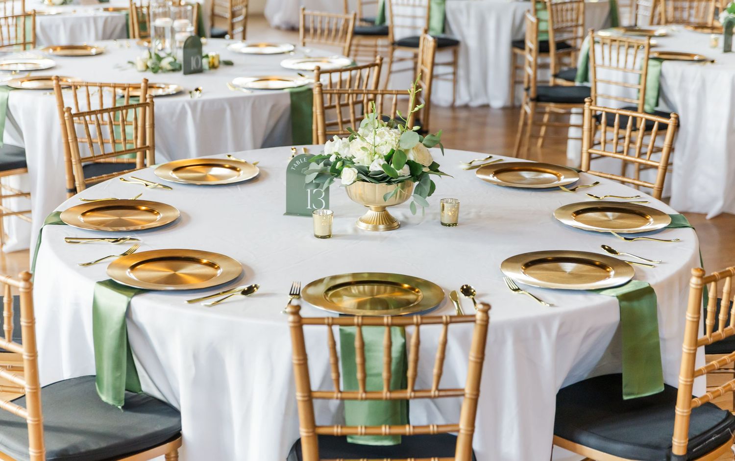 An elegant wedding reception table with gold chargers, chiavari chairs, and sage green accents on crisp white linens.