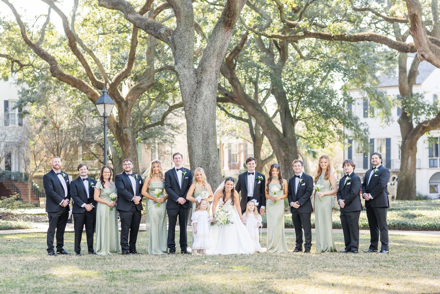 Wedding party in formal attire stands in a line under oak trees on a sunny afternoon.