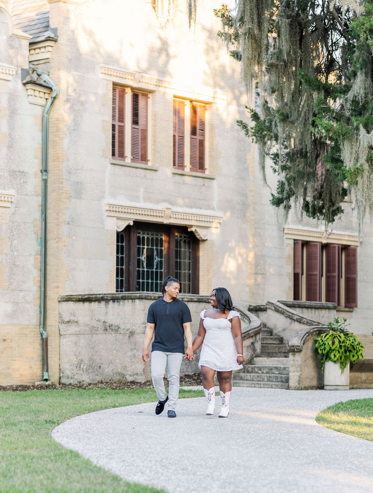 A couple walks along a curved path in front of a historic stone building with Spanish moss hanging from trees.